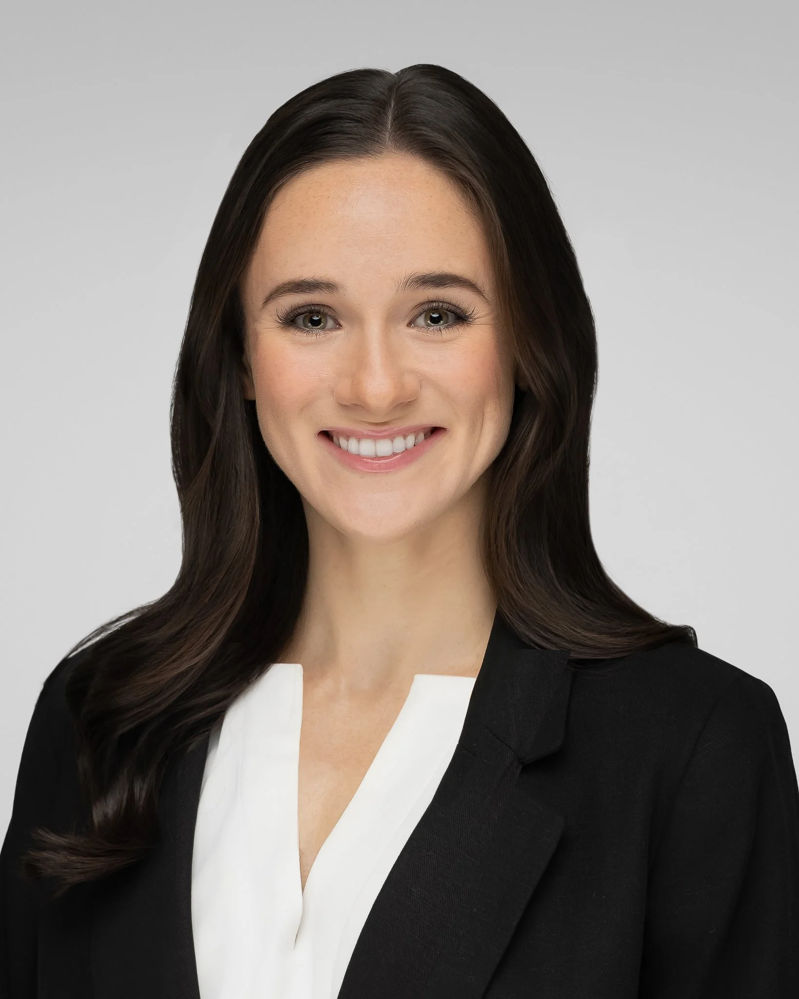 Close-up of a woman with long brown hair, wearing a black and white professional outfit, smiling against a plain gray background.