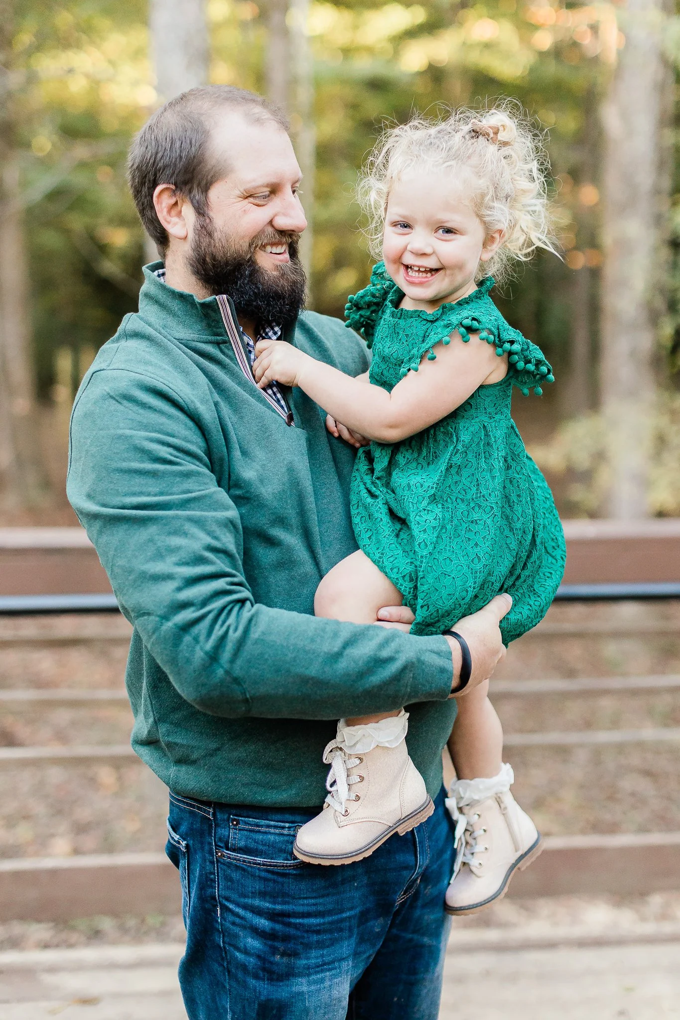 A man holding a smiling toddler in a green dress, outdoors in a park.