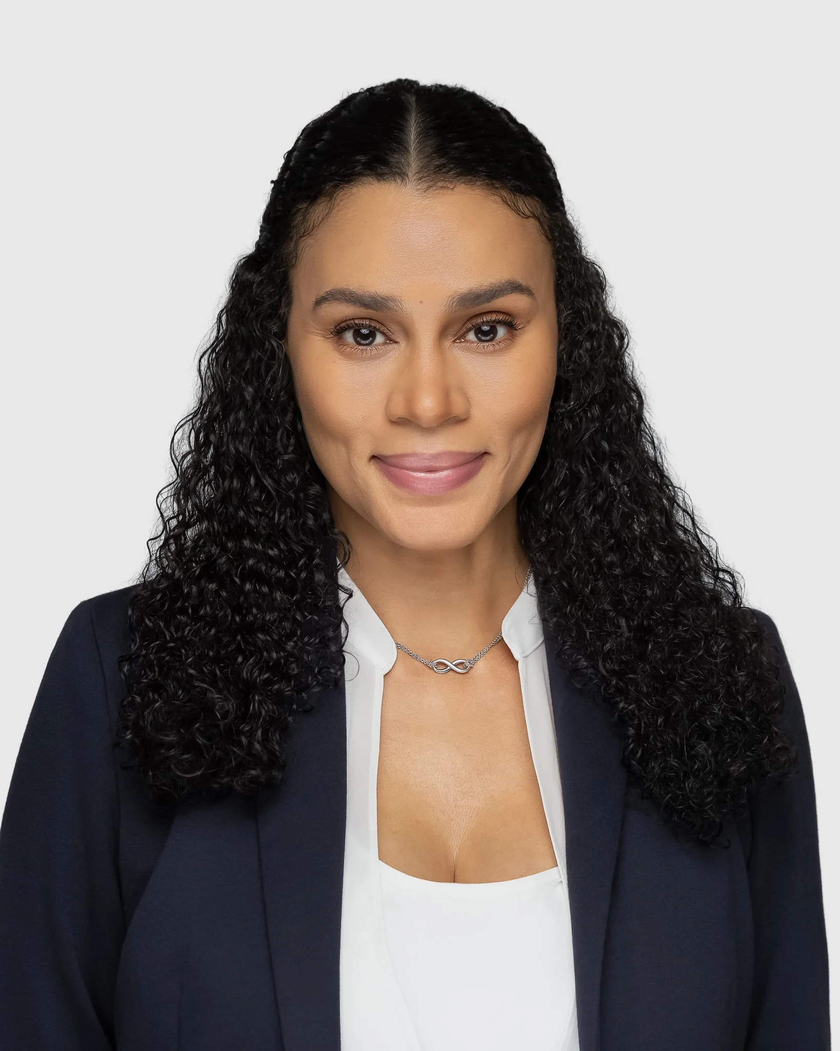 Close-up of a woman with curly dark hair, wearing a navy blazer over a white blouse with a scooped neckline, and a silver infinity necklace, smiling softly with bare skin.