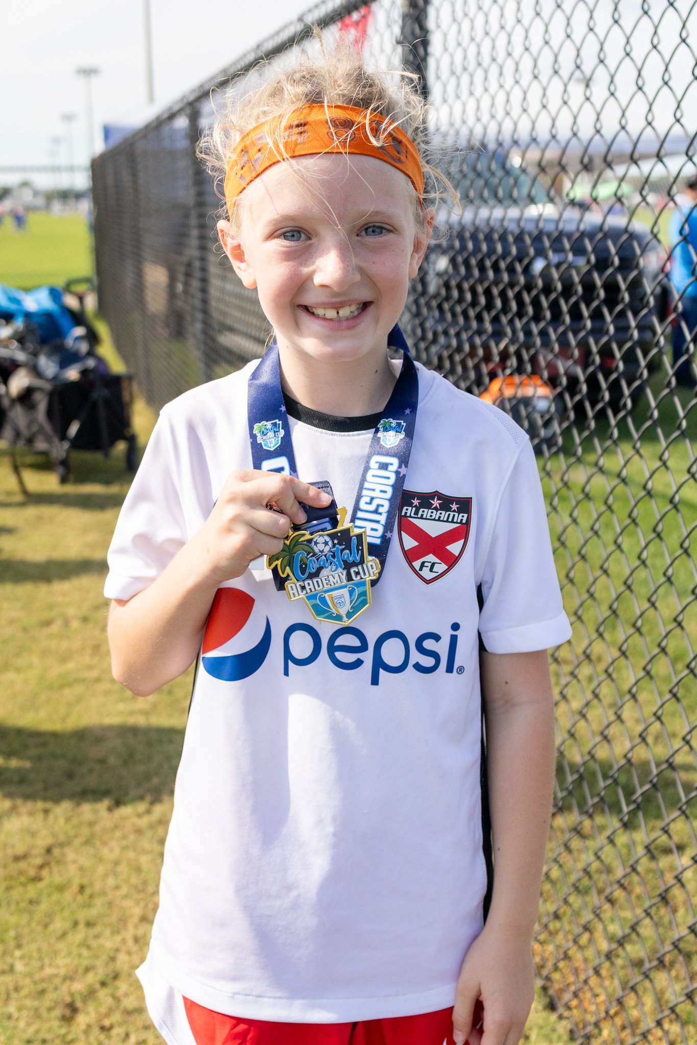 A young girl with blonde hair, an orange headband, and blue eyes, smiling while holding a medal. She is wearing a white soccer jersey. She is standing outdoors on a grassy area near a black chain-link fence.