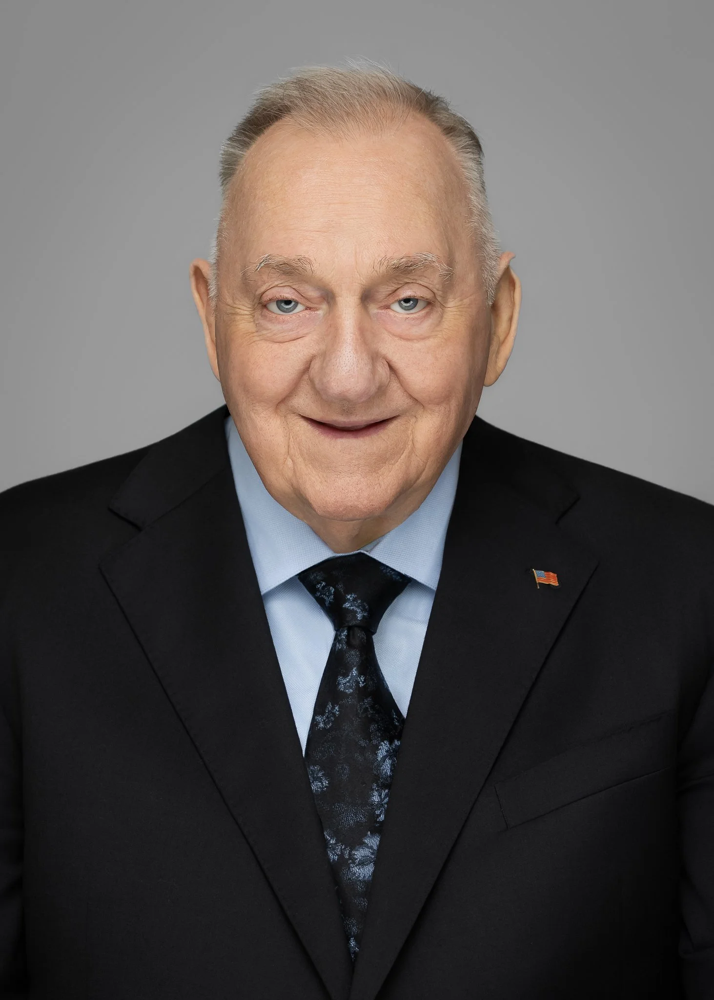 Portrait of an elderly man with light skin, blue eyes, and gray hair, wearing a black suit, light blue shirt, dark patterned tie, and a small American flag pin.