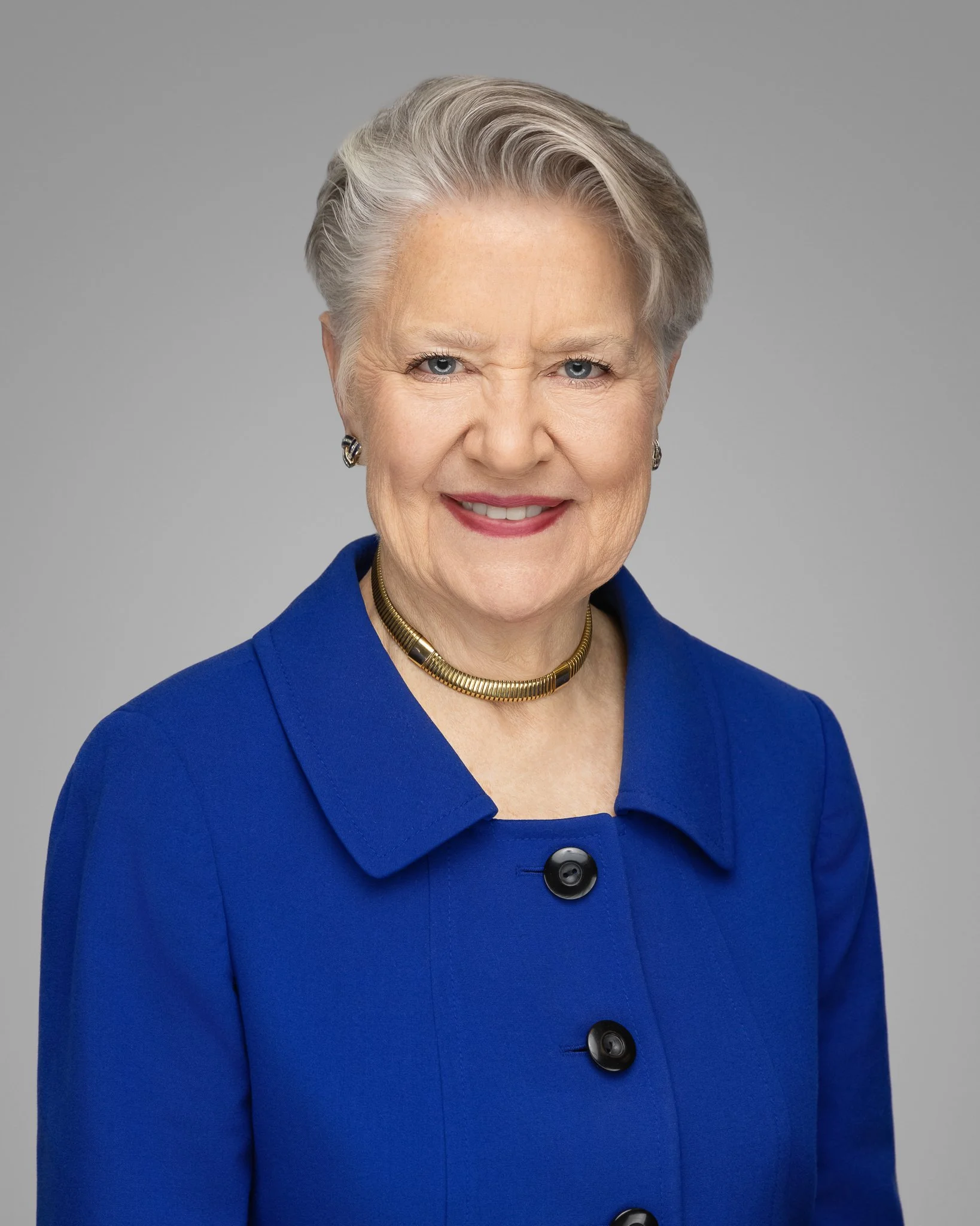 Close-up of an elderly woman with short gray hair, wearing a royal blue coat with large black buttons, a gold necklace, and earrings, smiling against a plain gray background.