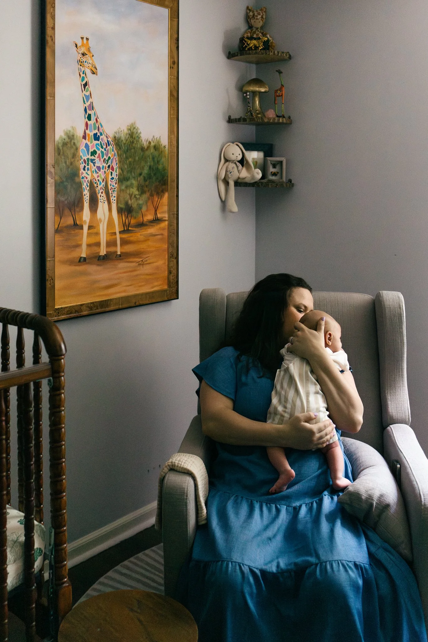 A woman with dark hair sitting in a beige armchair, breastfeeding a baby dressed in a striped outfit, in a cozy living room with a large canvas painting of a giraffe on the wall and decorative shelves with stuffed animals and figurines.