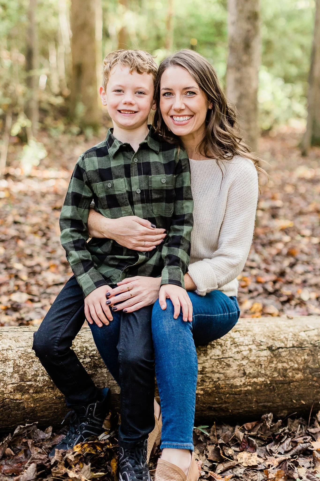A woman and boy sitting on a log in a forest, wearing casual clothes, surrounded by autumn leaves.