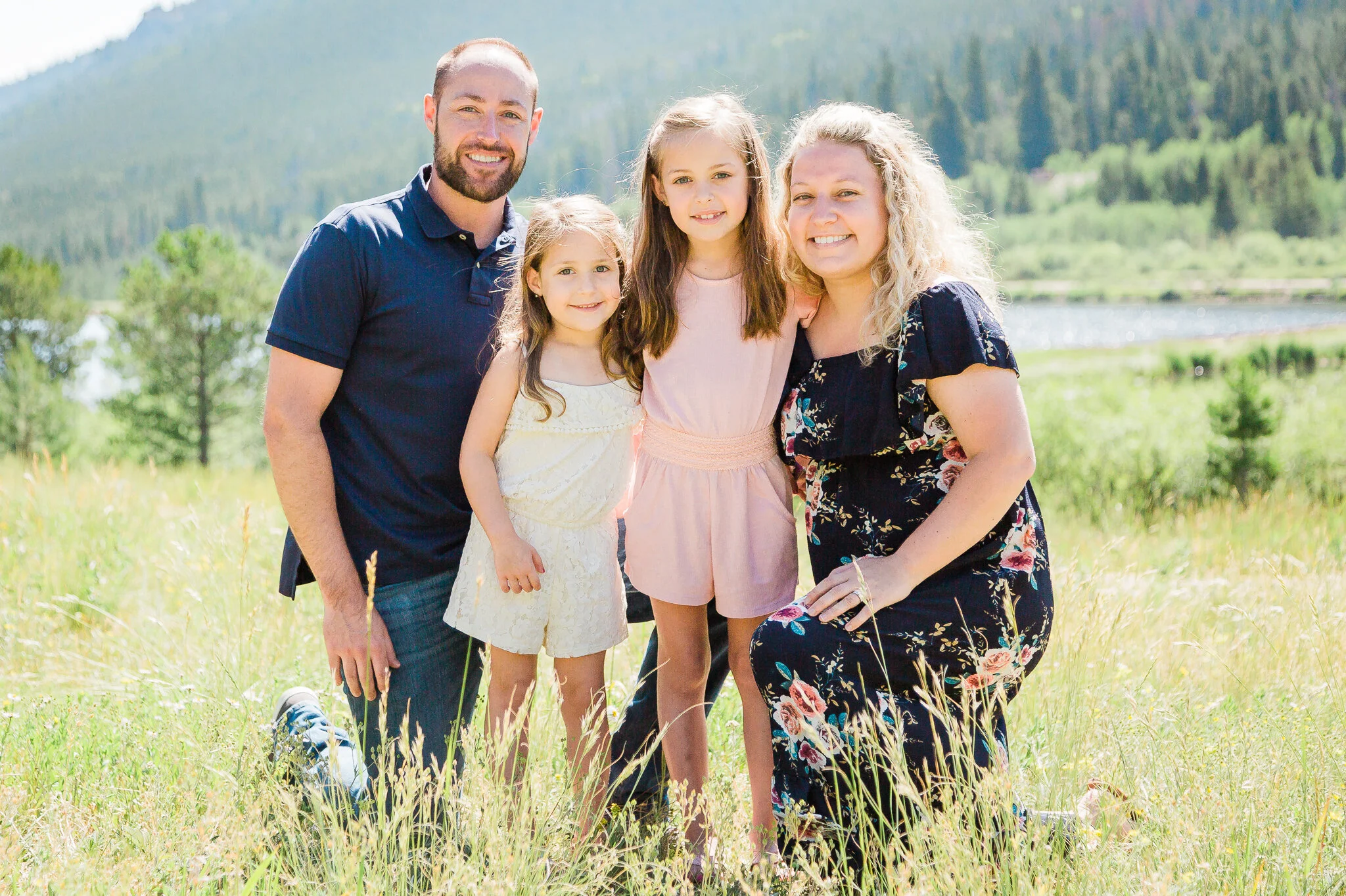 Family of four posing outdoors in a grassy field with mountains in the background.