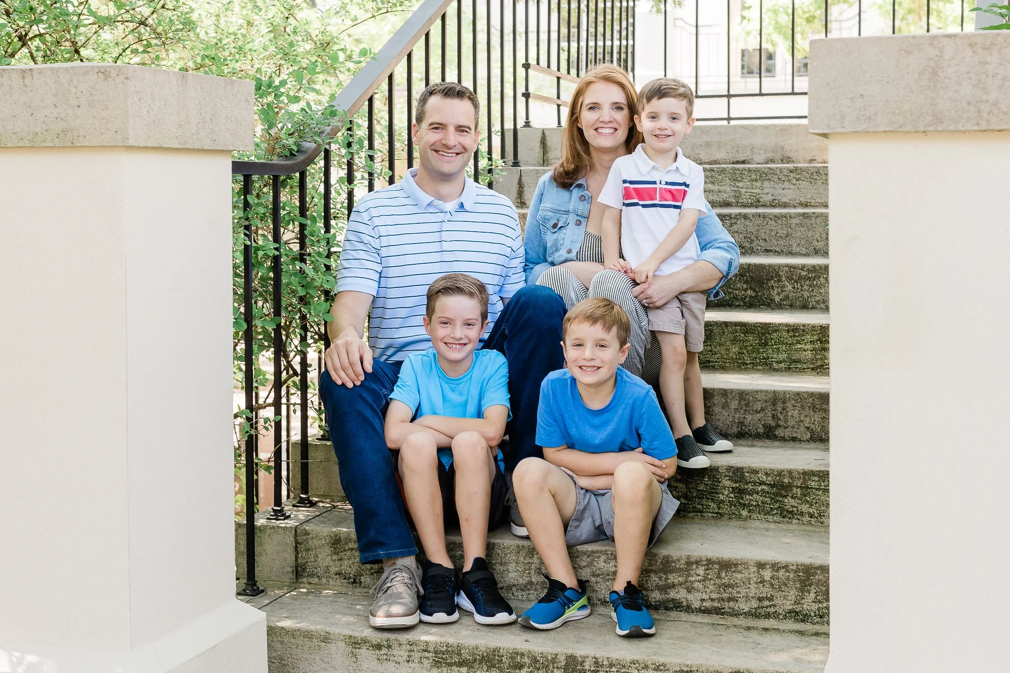 Family of five sitting on outdoor stairs, smiling, with greenery in the background.