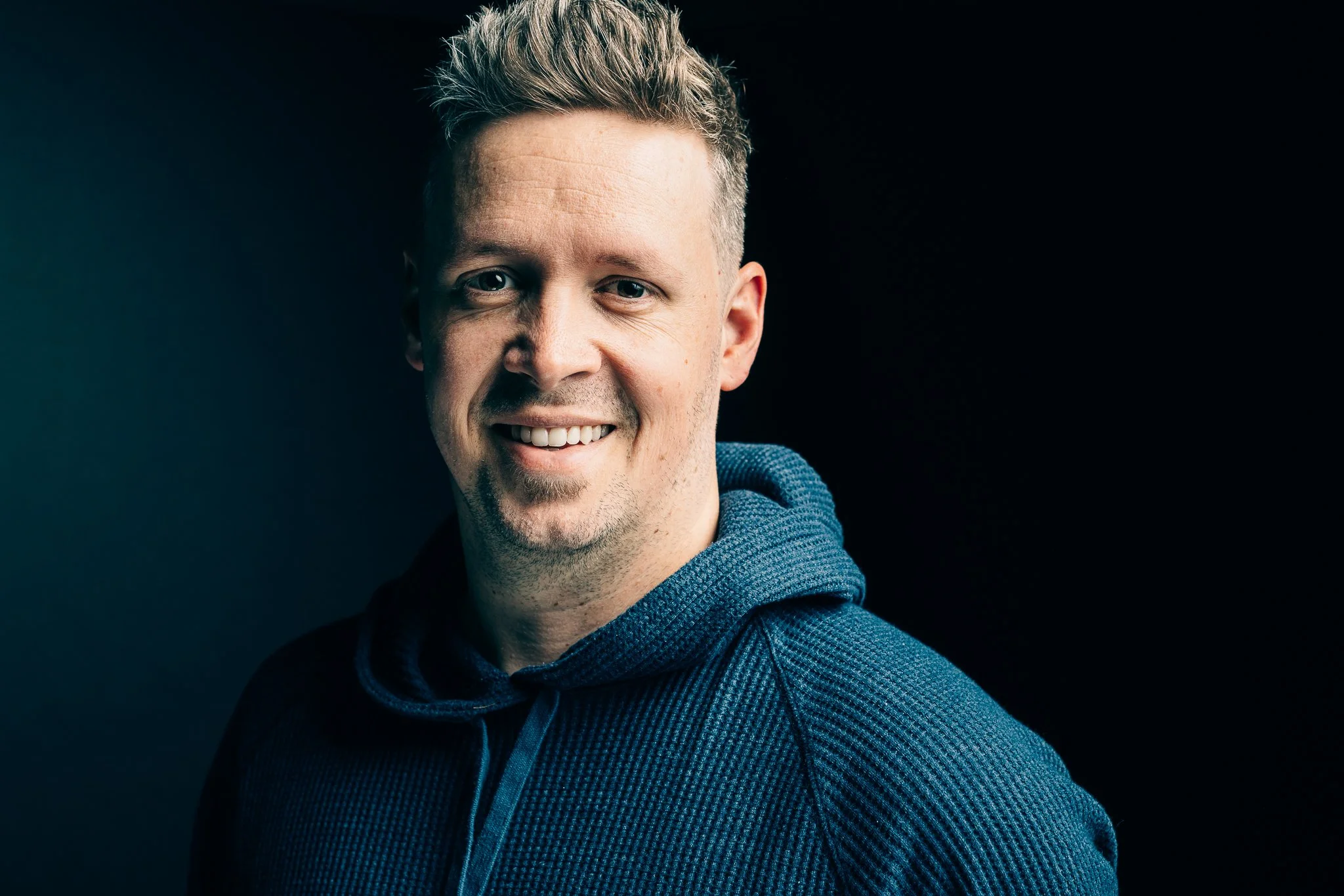A portrait of Seth Allen of Seth James Photography with short, styled blonde hair wearing a dark blue hoodie, standing against a dark background.