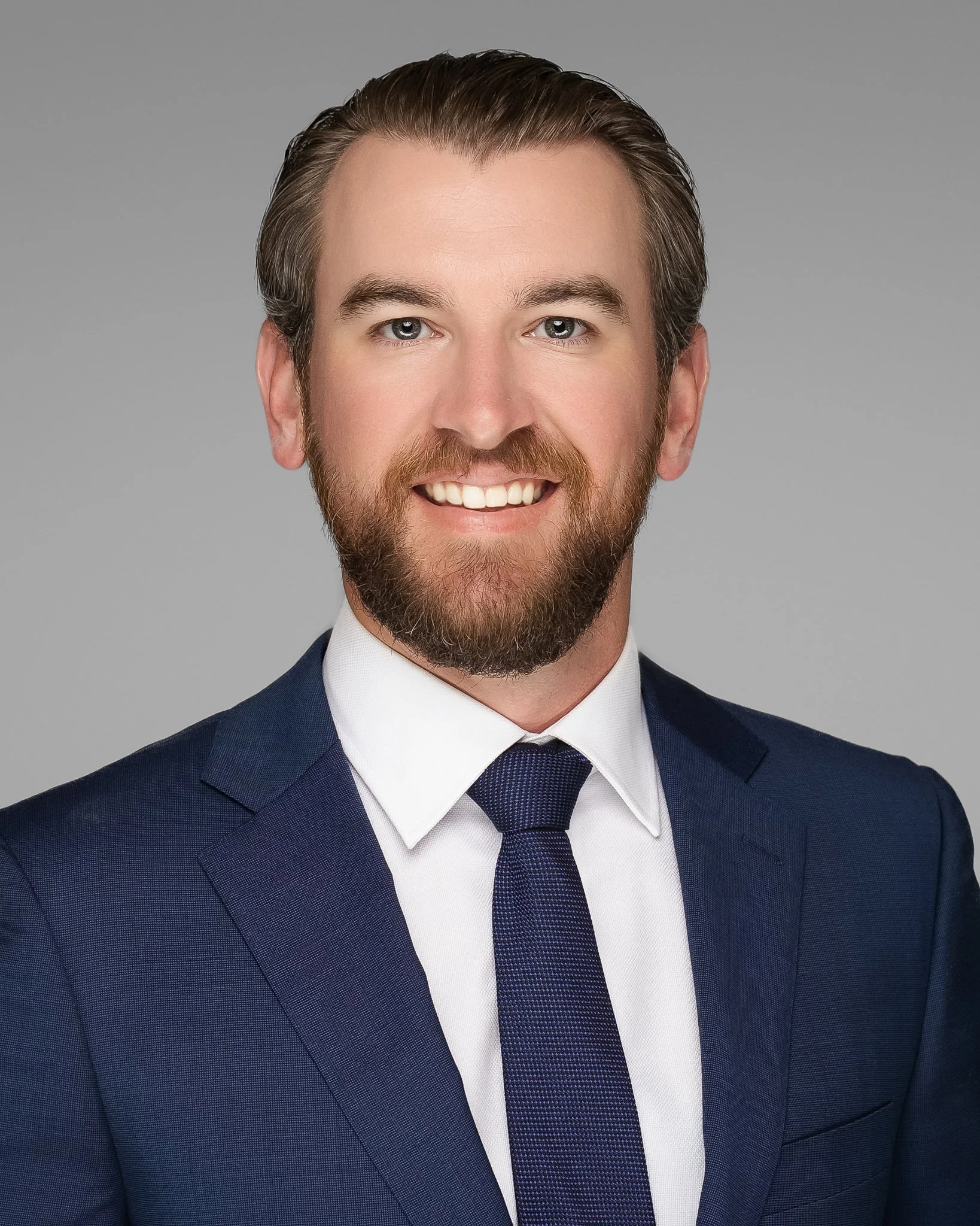 A professional headshot of a smiling man with light skin, brown hair, a beard, wearing a navy suit, white shirt, and navy tie, against a gray background.