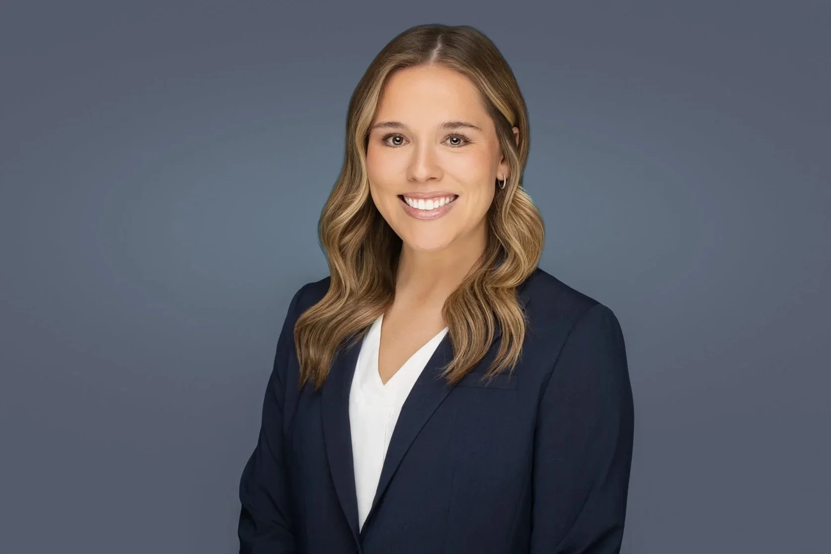 A professional headshot of a woman with wavy light brown hair, smiling, dressed in a dark blazer over a white top, against a plain gray background.