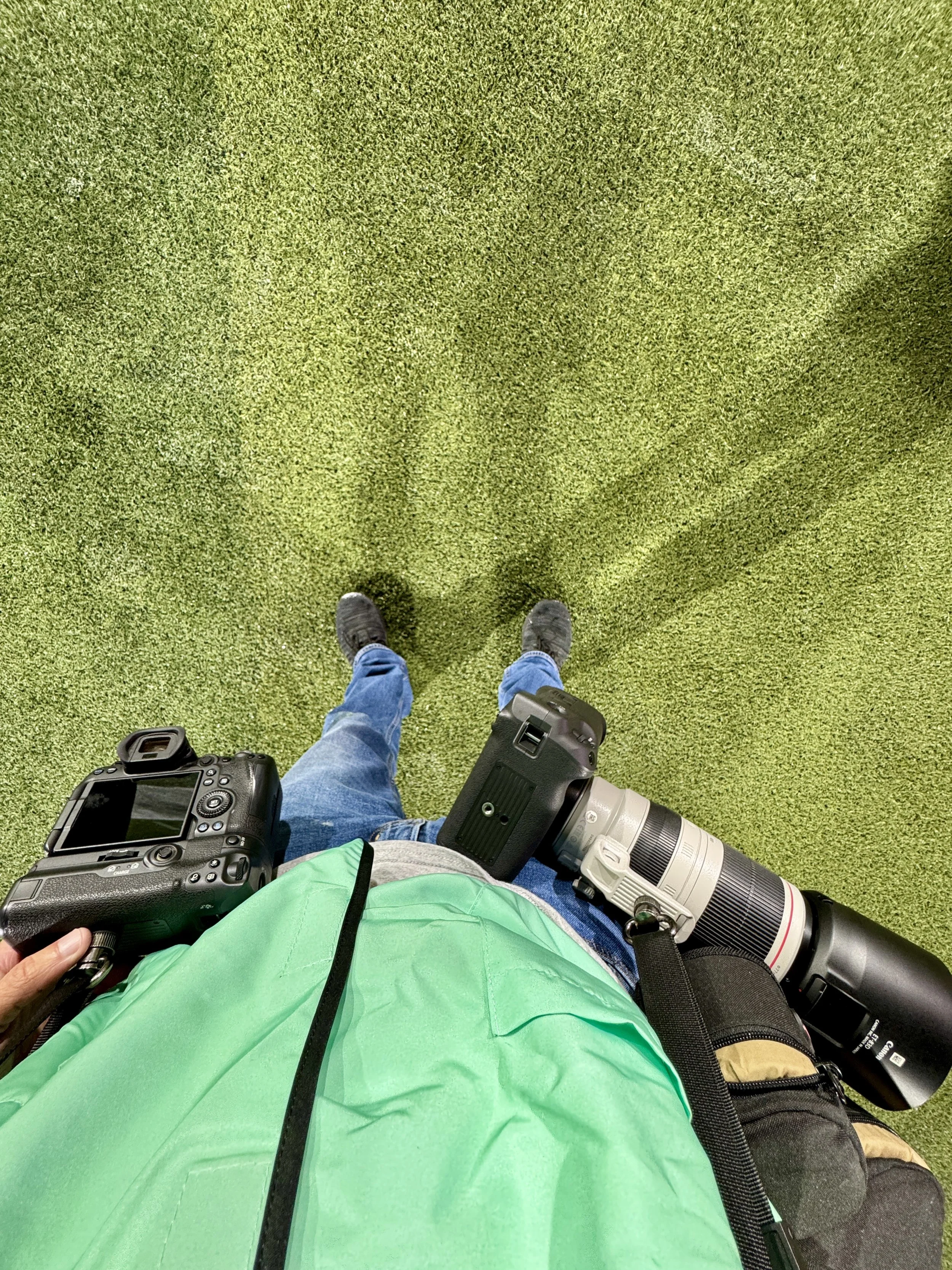 Overhead view of a sports photographer in Birmingham Alabama standing on green outdoor turf, holding a camera and a camera lens with a strap, wearing blue jeans and gray shoes, with a green backpack or jacket.