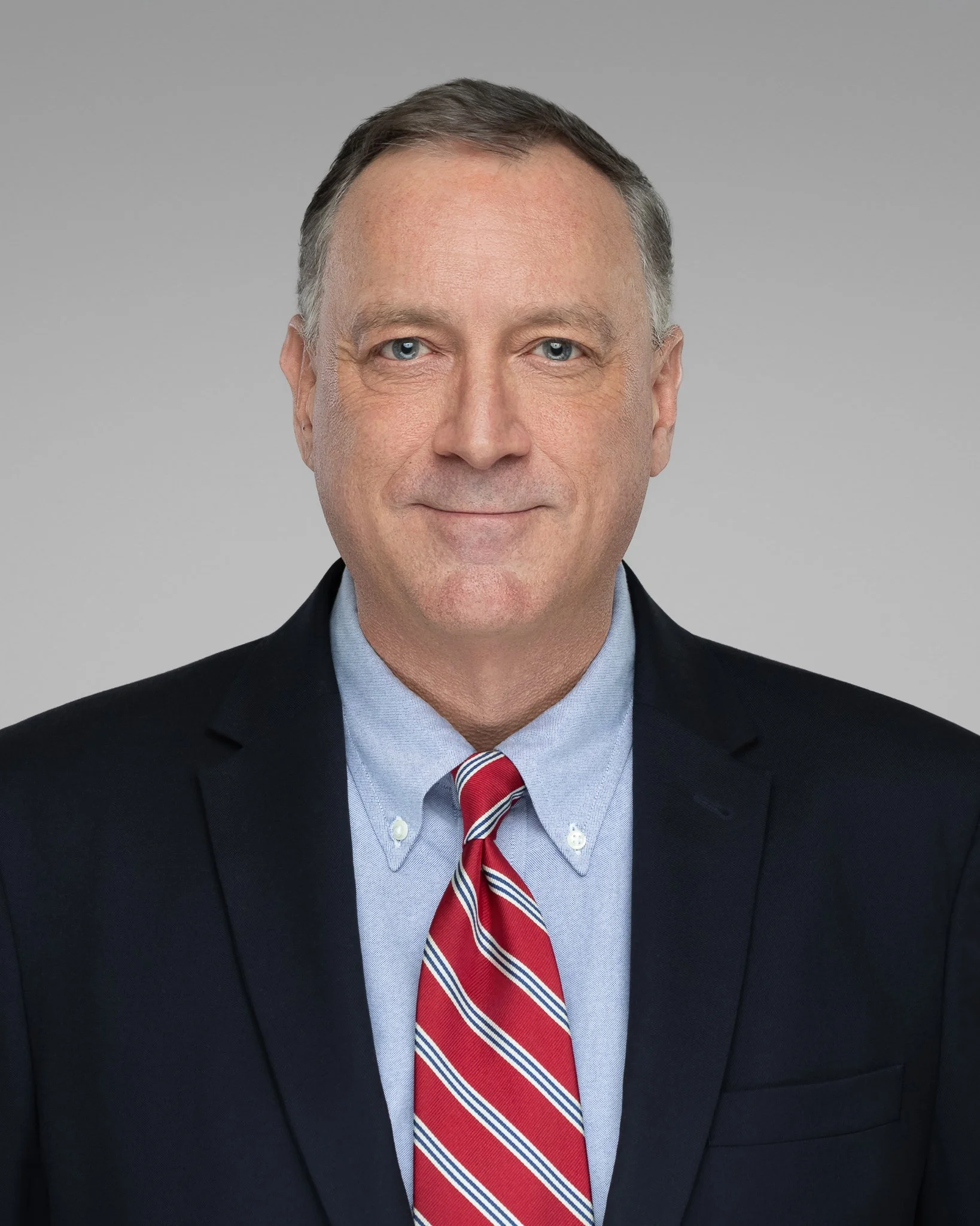 A middle-aged man dressed in a black suit, light blue shirt, and a red, white, and blue striped tie, standing against a plain gray background.