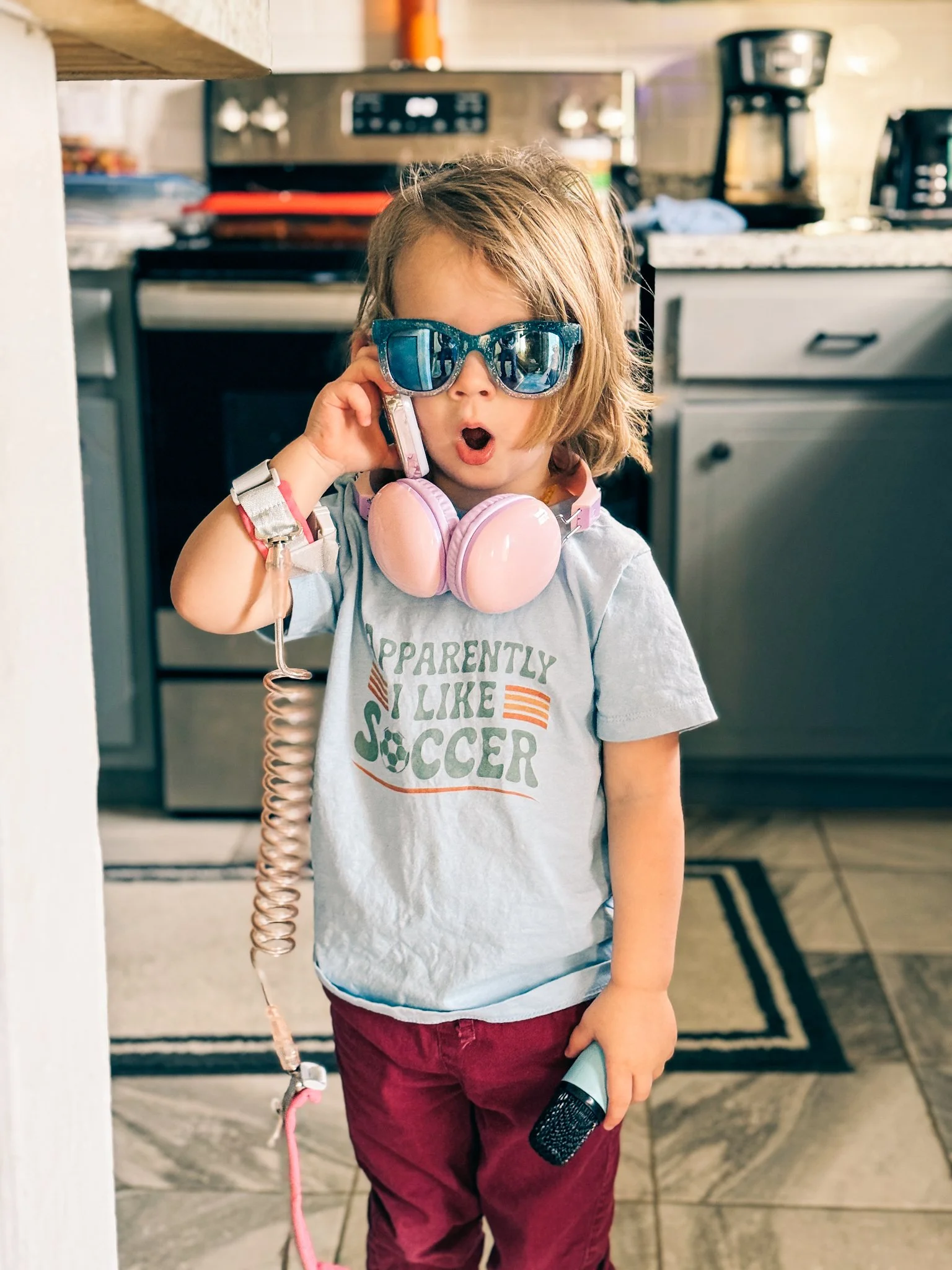 A young child with shaggy hair wearing blue sunglasses and pink headphones around her neck, holding a toy microphone, standing in a kitchen.