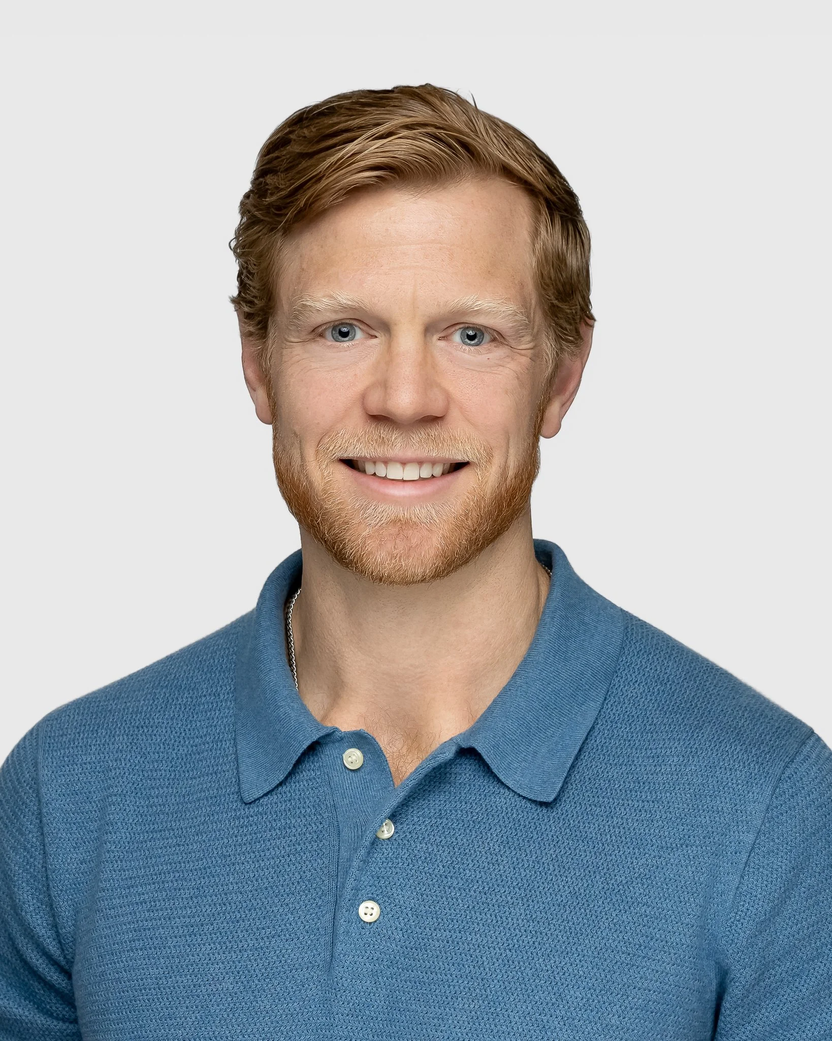Close-up of a smiling man with red hair and beard, wearing a blue button-up shirt, against a plain gray background.