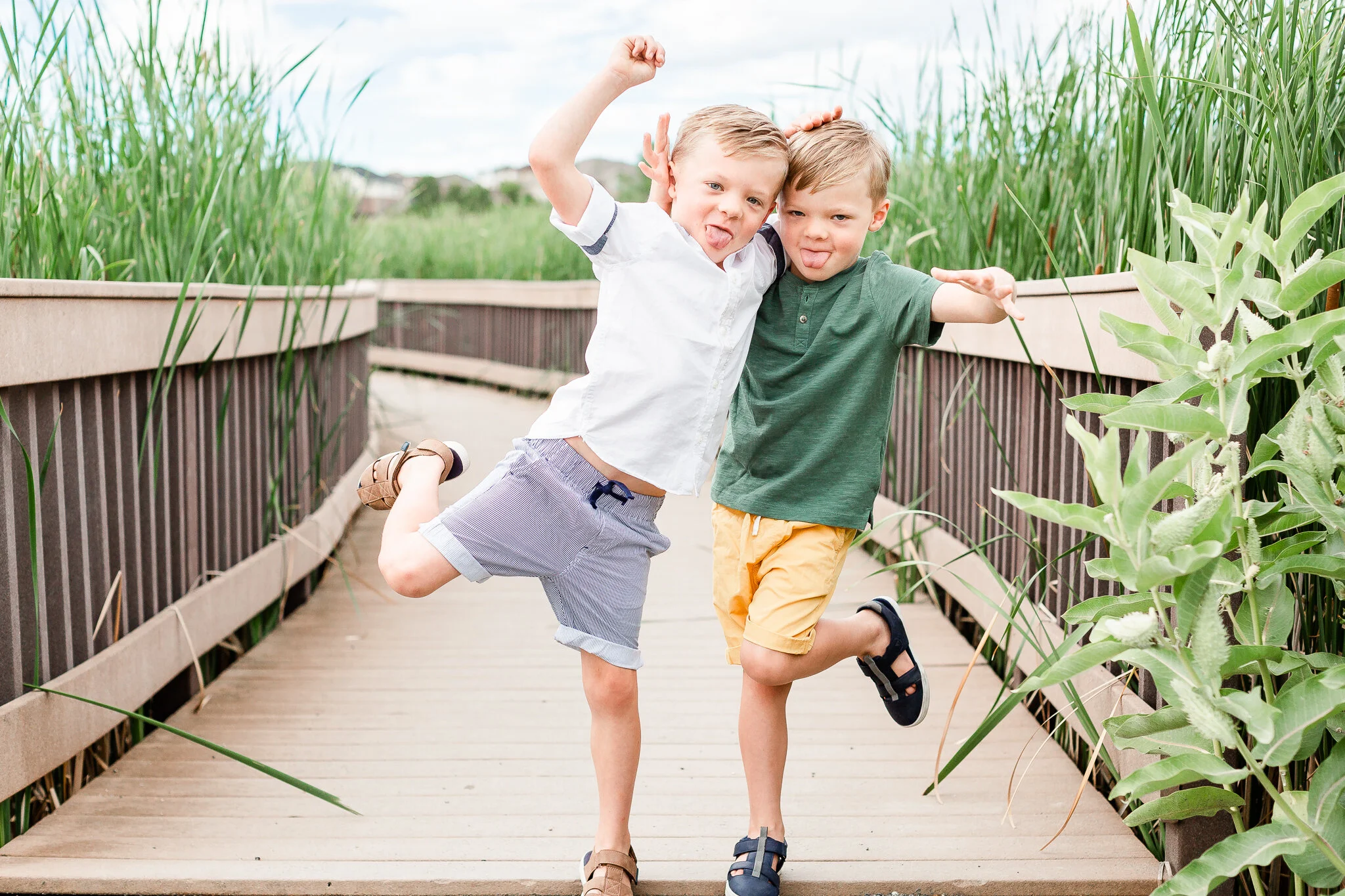 Two young boys making funny faces and poses on a wooden walkway surrounded by tall grass.