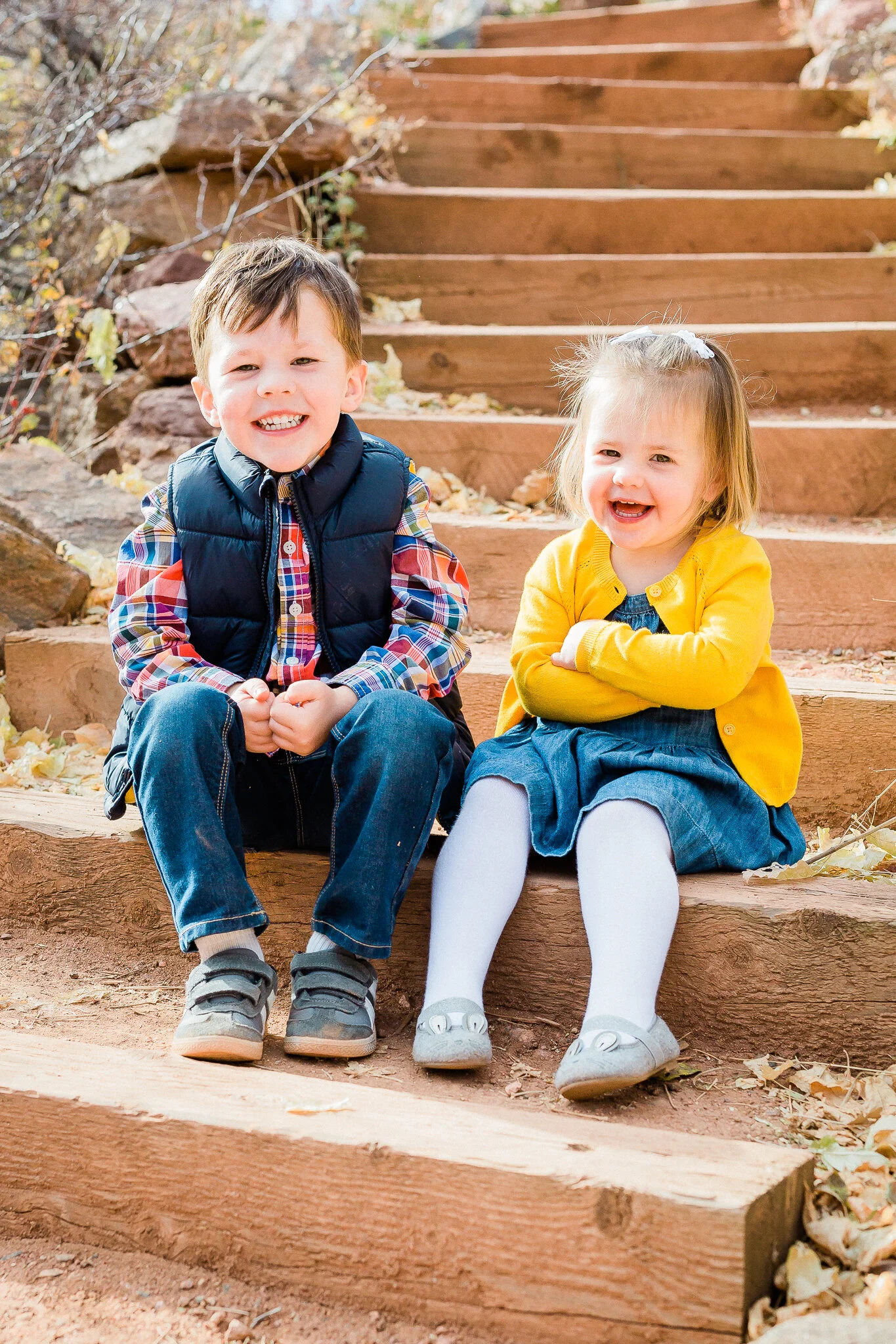Two children sitting on wooden steps outdoors, smiling. The boy wears a black vest and plaid shirt; the girl wears a yellow cardigan and denim dress. Autumn leaves are scattered around.