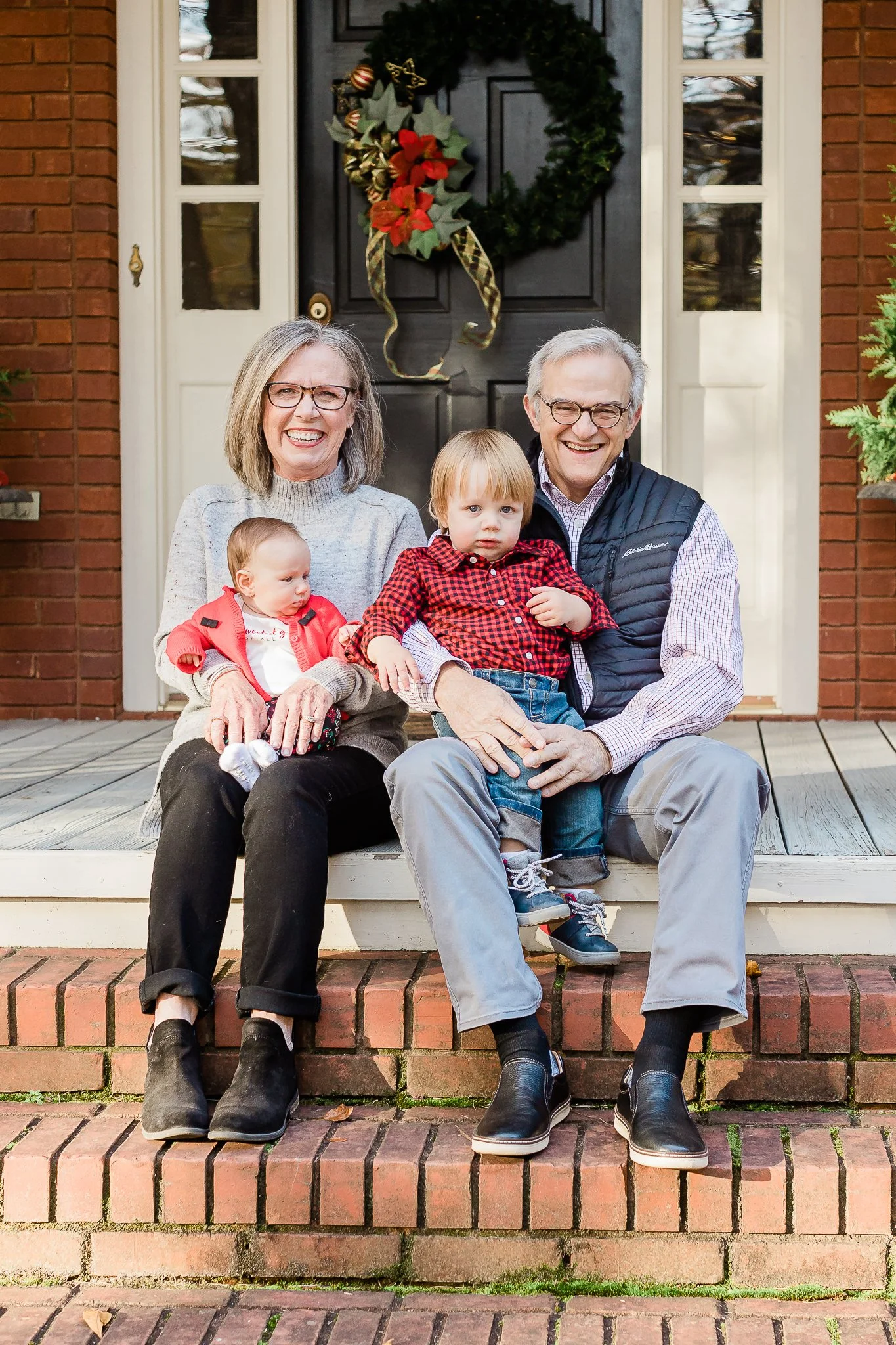 Older couple sitting on brick steps with two young children, Christmas wreath on the door behind them.