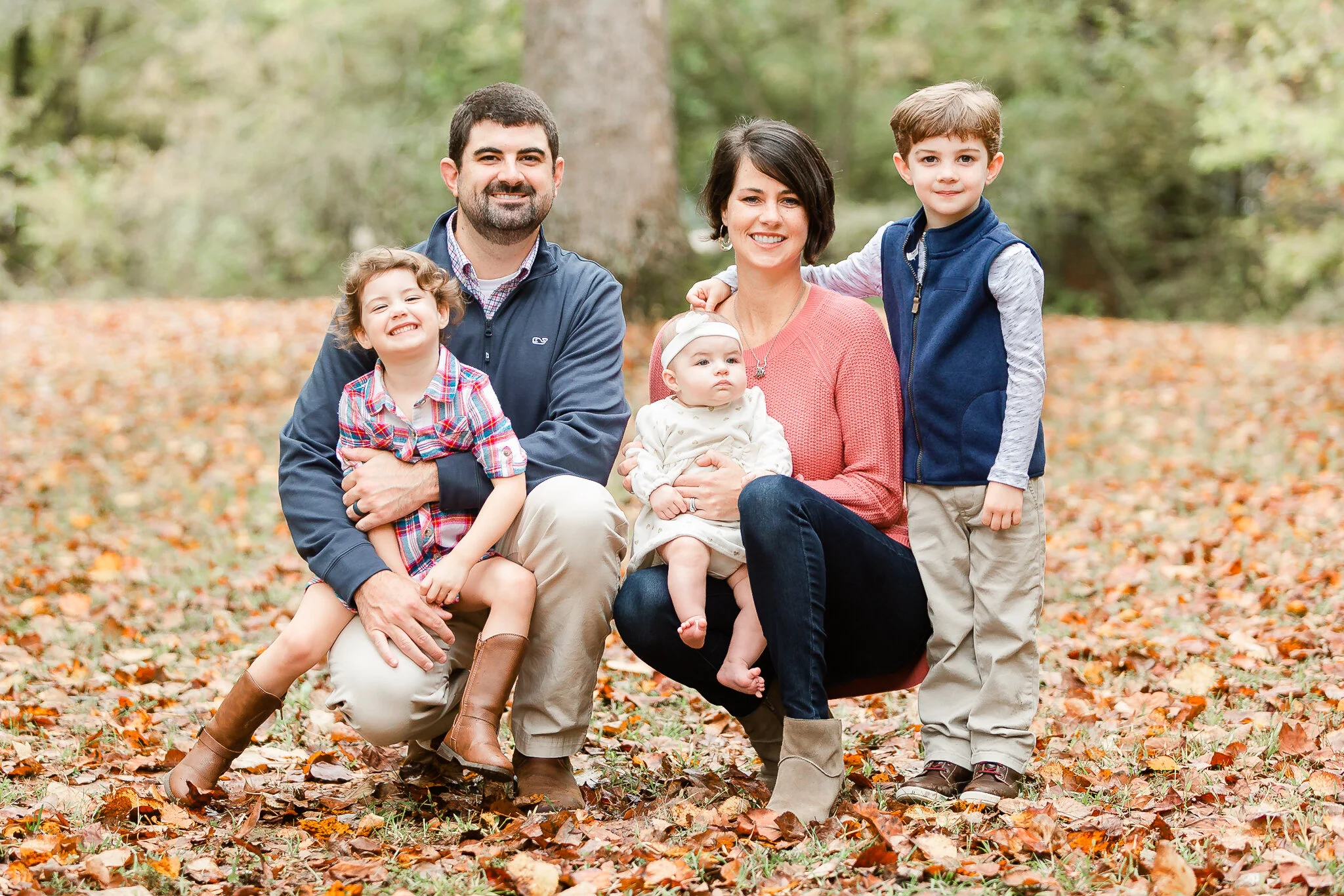 A family of five posing outdoors during autumn, surrounded by fallen leaves. The father is crouching, holding a smiling child, while the mother is sitting with an infant on her lap. Another child stands beside them. The family is dressed in casual fa