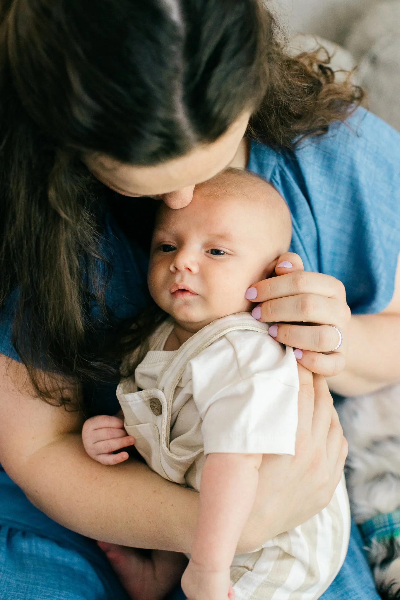 An adult woman with long dark hair holding a baby with light coloring and a bald head. The woman is whispering or kissing the baby, who is looking slightly to the side. The woman wears a blue top, and the baby wears a beige and white striped outfit.