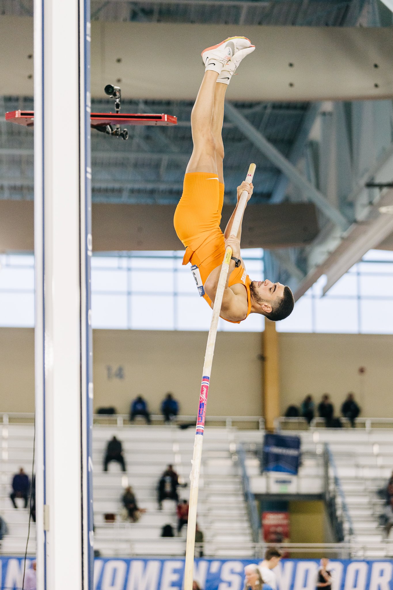 Athlete performing a pole vault indoors, wearing an orange uniform, with spectators in the background.