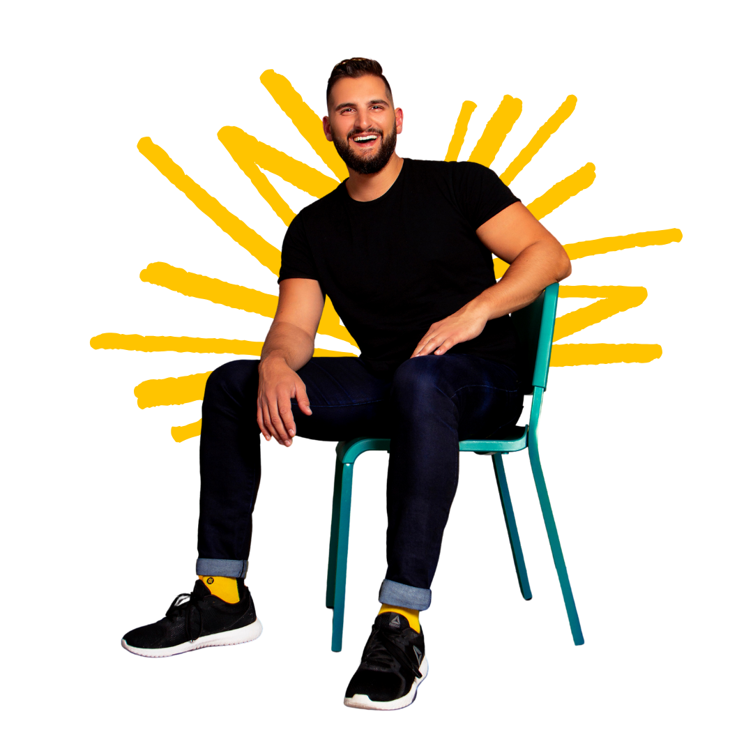 A smiling man with a beard wearing a black t-shirt, jeans, yellow socks, and black sneakers, sitting on a teal chair against a colorful, abstract background.