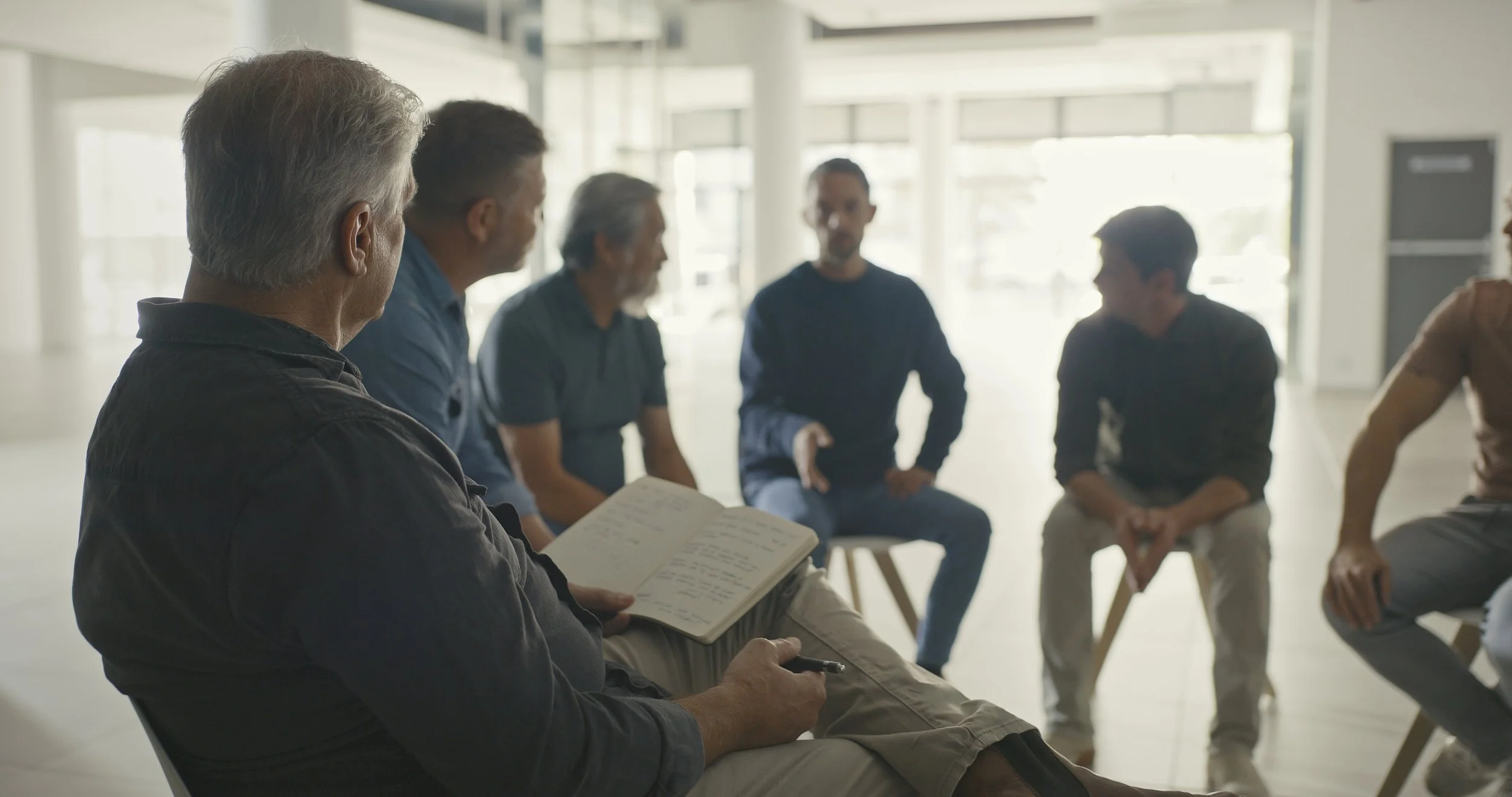 Group of five men sitting in a circle in a brightly lit room with large windows, engaged in discussion or meeting.