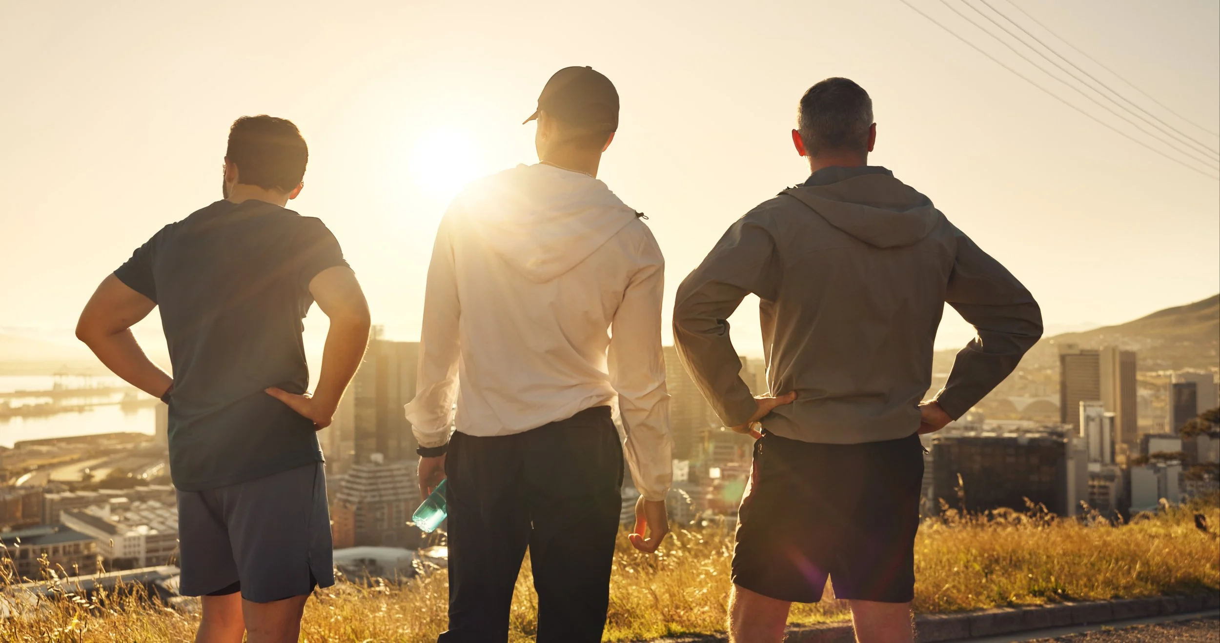 Three men stand on a hill overlooking a city at sunset, facing away from the camera.
