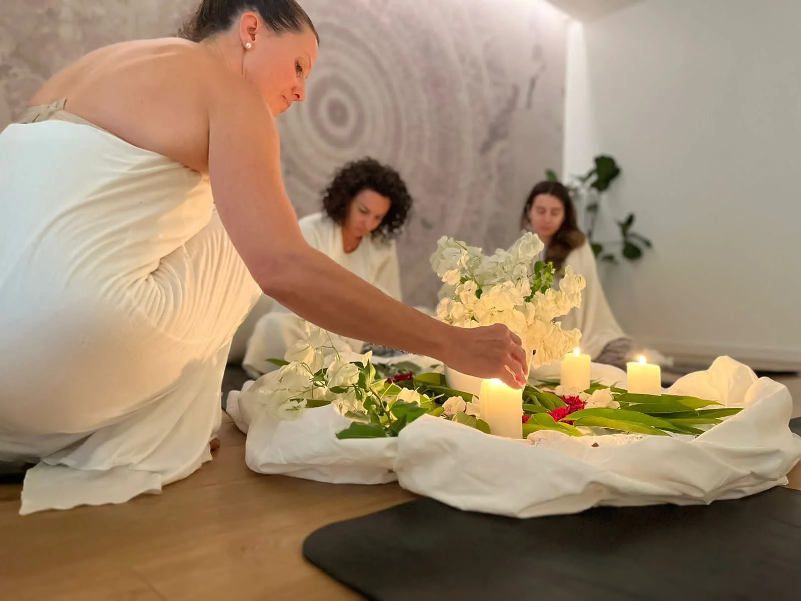 A woman lighting candles during a meditation or spiritual ceremony with two other women seated in the background, all dressed in white, in a peaceful indoor setting.