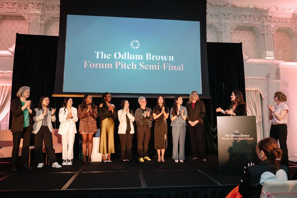 A group of women stand across a stage applauding. The screen behind them reads: The Odlum Brown Forum Pitch Semi-Final