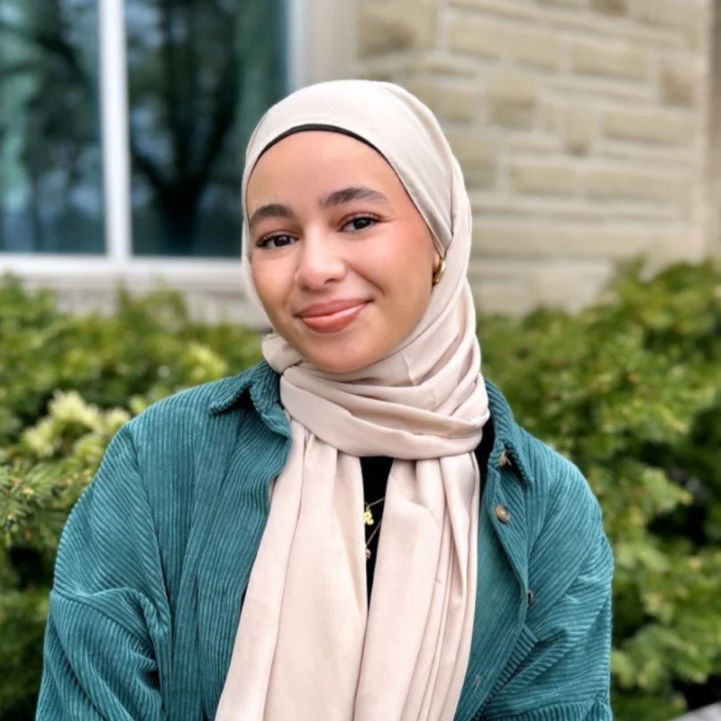 A woman in a long headscarf and blue corduroy jacket smiles in front of a building with windows and shrubs