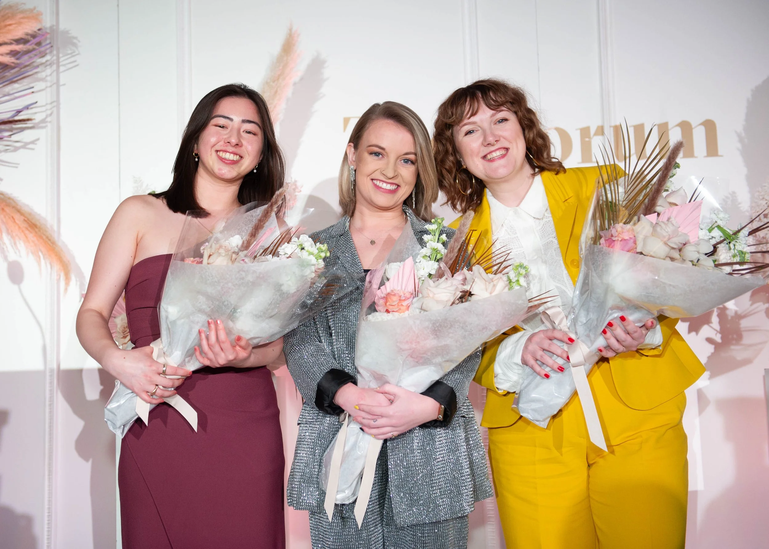 Three women stand on stage holding bouquets