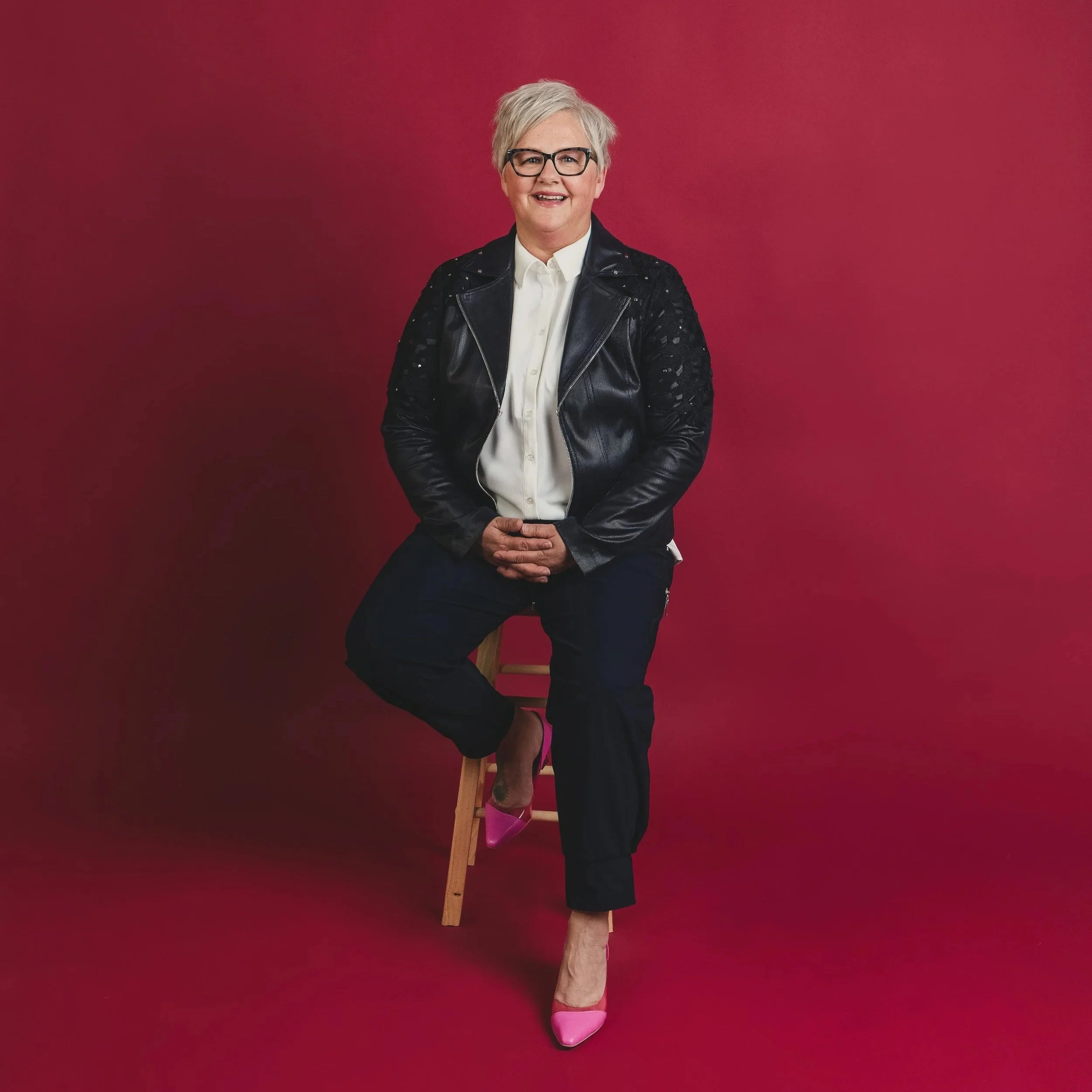 Woman with short white hair and dark framed glasses, wearing a black leather jacket over white shirt, black pants, and hot pink shoes, perches on a stool against a red background
