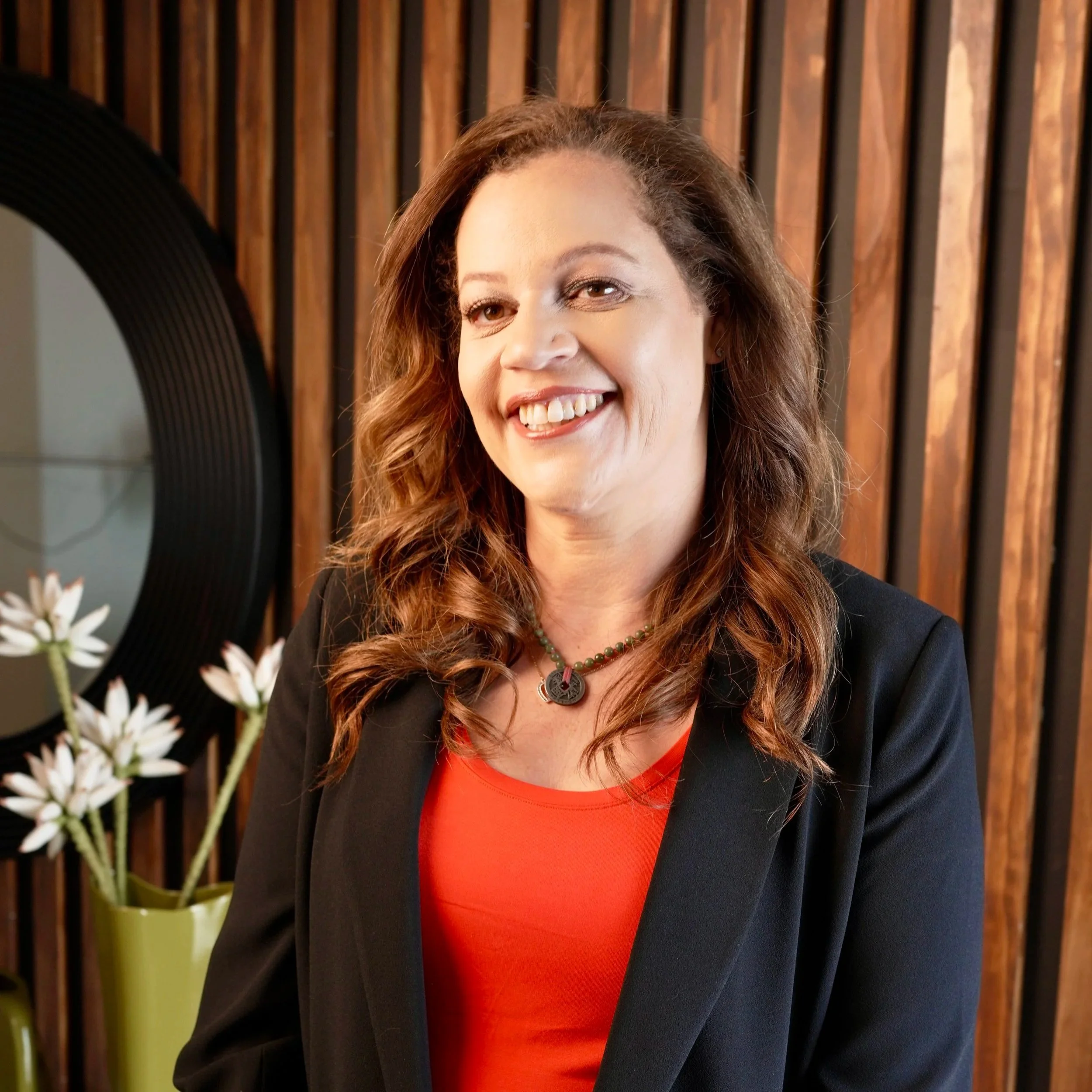 A smiling woman with brown curls is wearing a black blazer over an orange top.