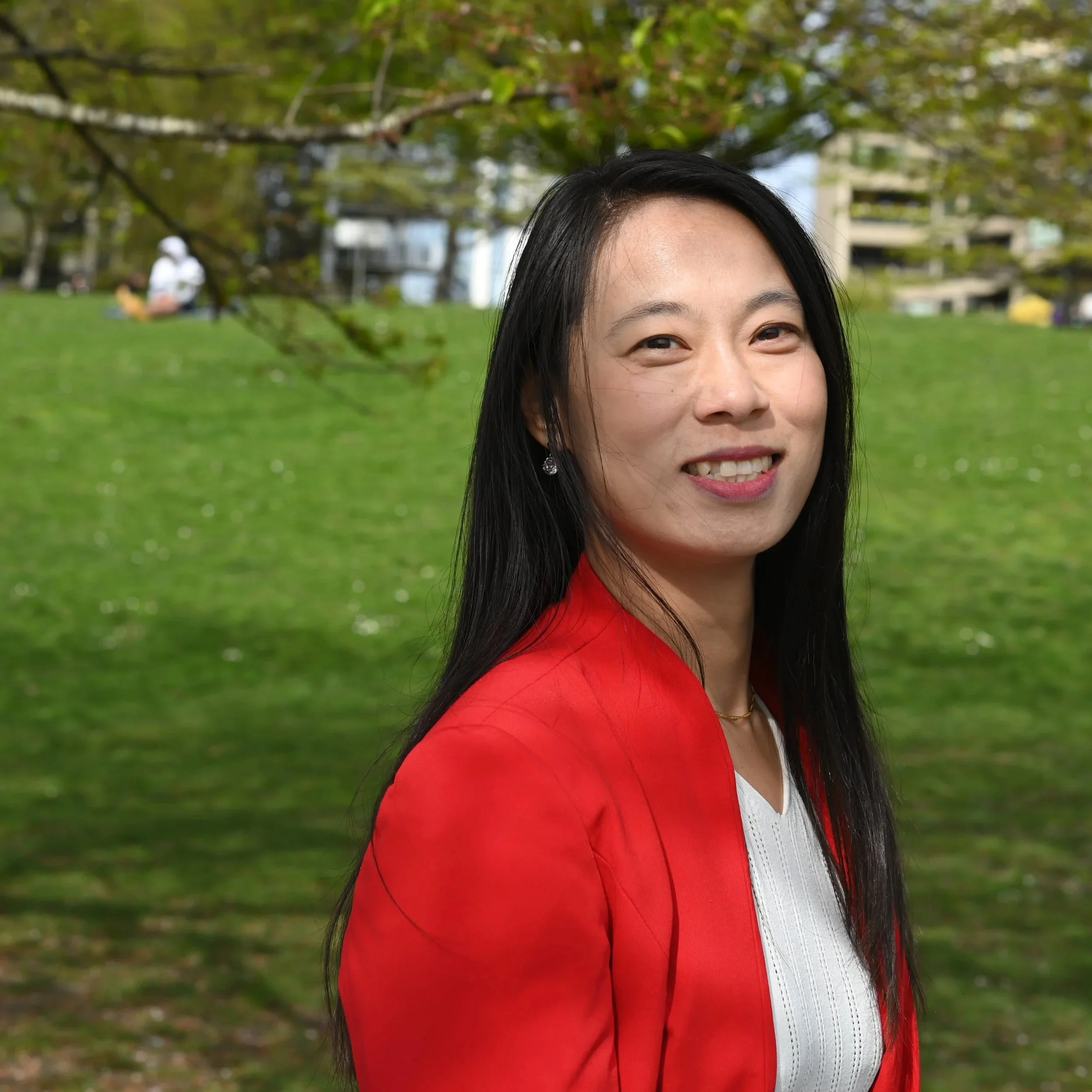 A woman with long black hair and an orange blazer sands in a park