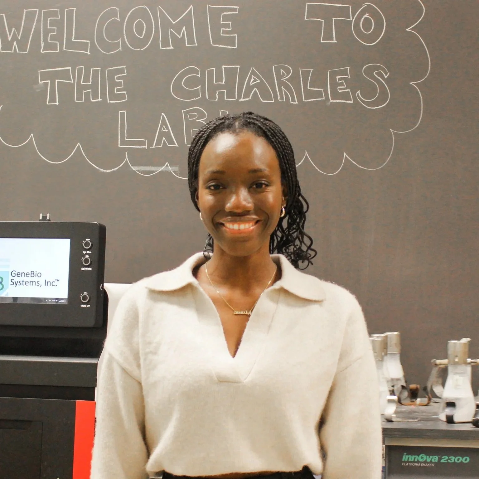 A woman in a white polo stands in front of a blackboard that reads Welcome to the Charles Lab