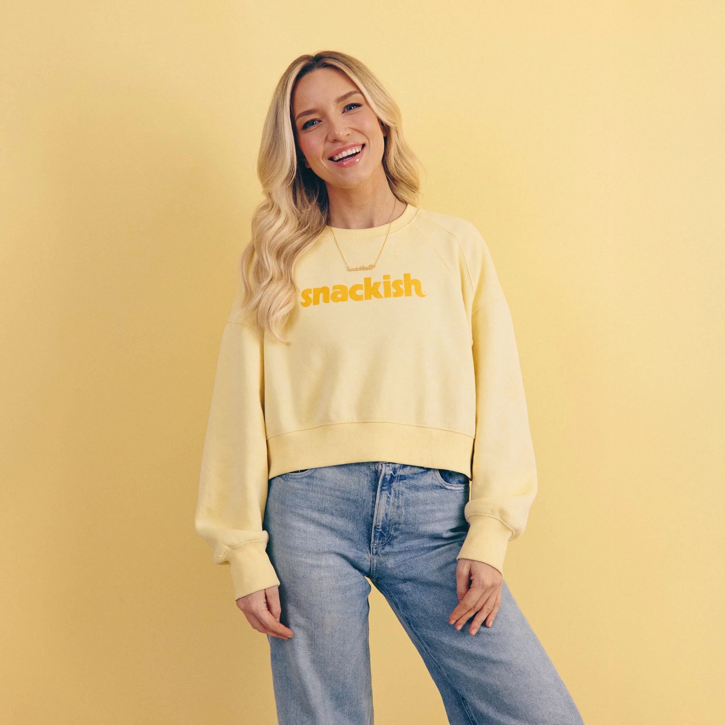 Smiling woman in a yellow sweatshirt that reads "snackish"