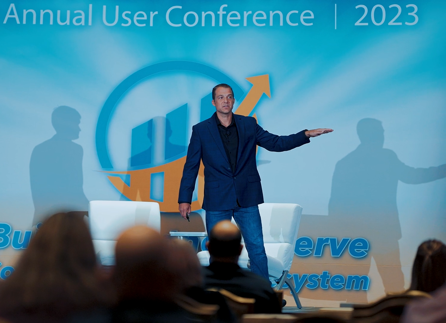 Man in a dark blazer speaking at an annual user conference, standing in front of a large screen with graphics and text about business growth and systems, 2023.