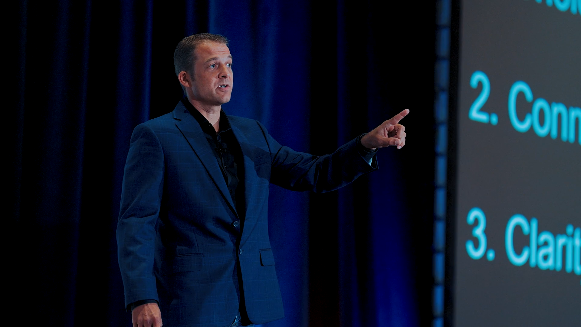 A man in a dark blue checkered suit giving a presentation, pointing at a slide with blue text on a large screen.