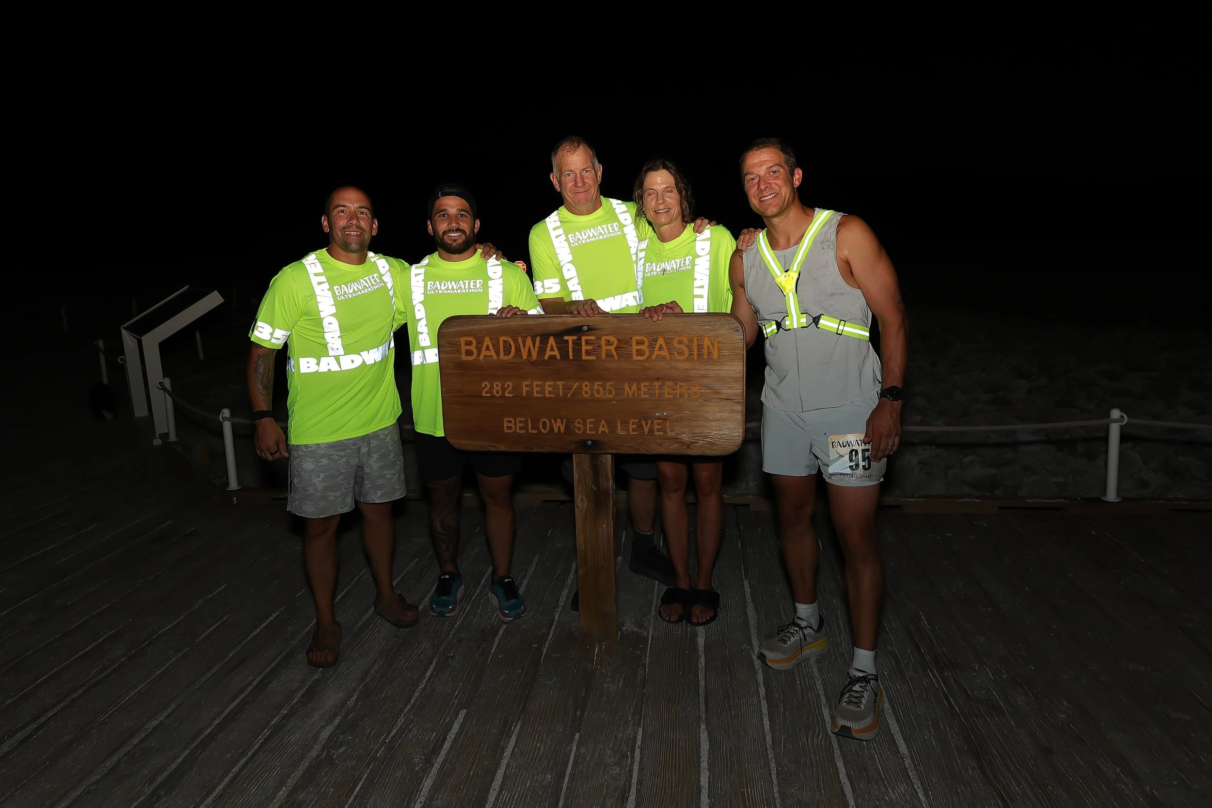 Group of five people standing behind a sign at Badwater Basin during nighttime; they are wearing bright yellow-green shirts and athletic gear, and the sign indicates they are 282 feet below sea level.