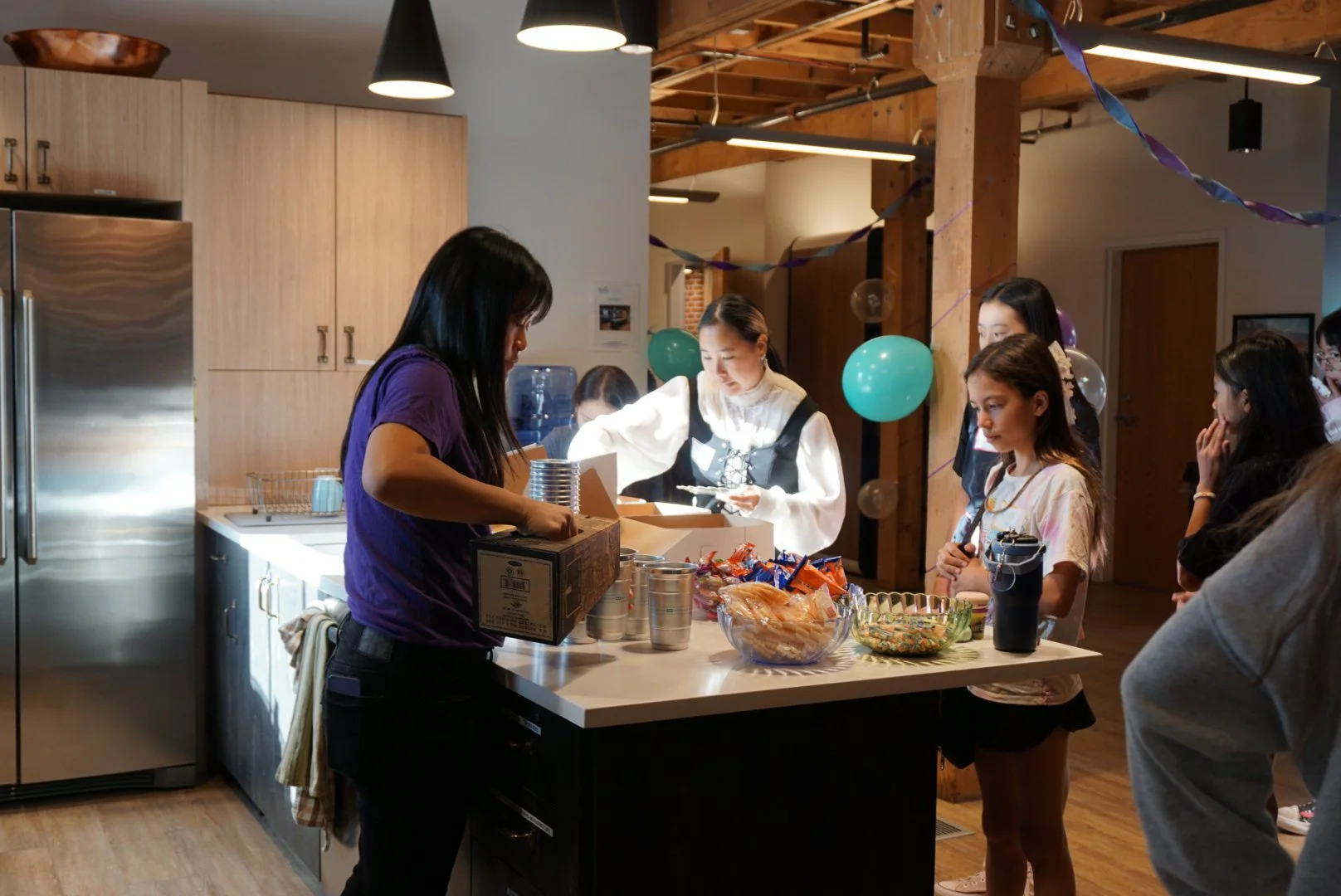  Woman in purple shirt in front of wooden cabinets and refrigerator pouring drink into aluminum cups serving breakfast with students looking and grabbing at snacks, wooden beams decorated with teal and purple balloons in background 