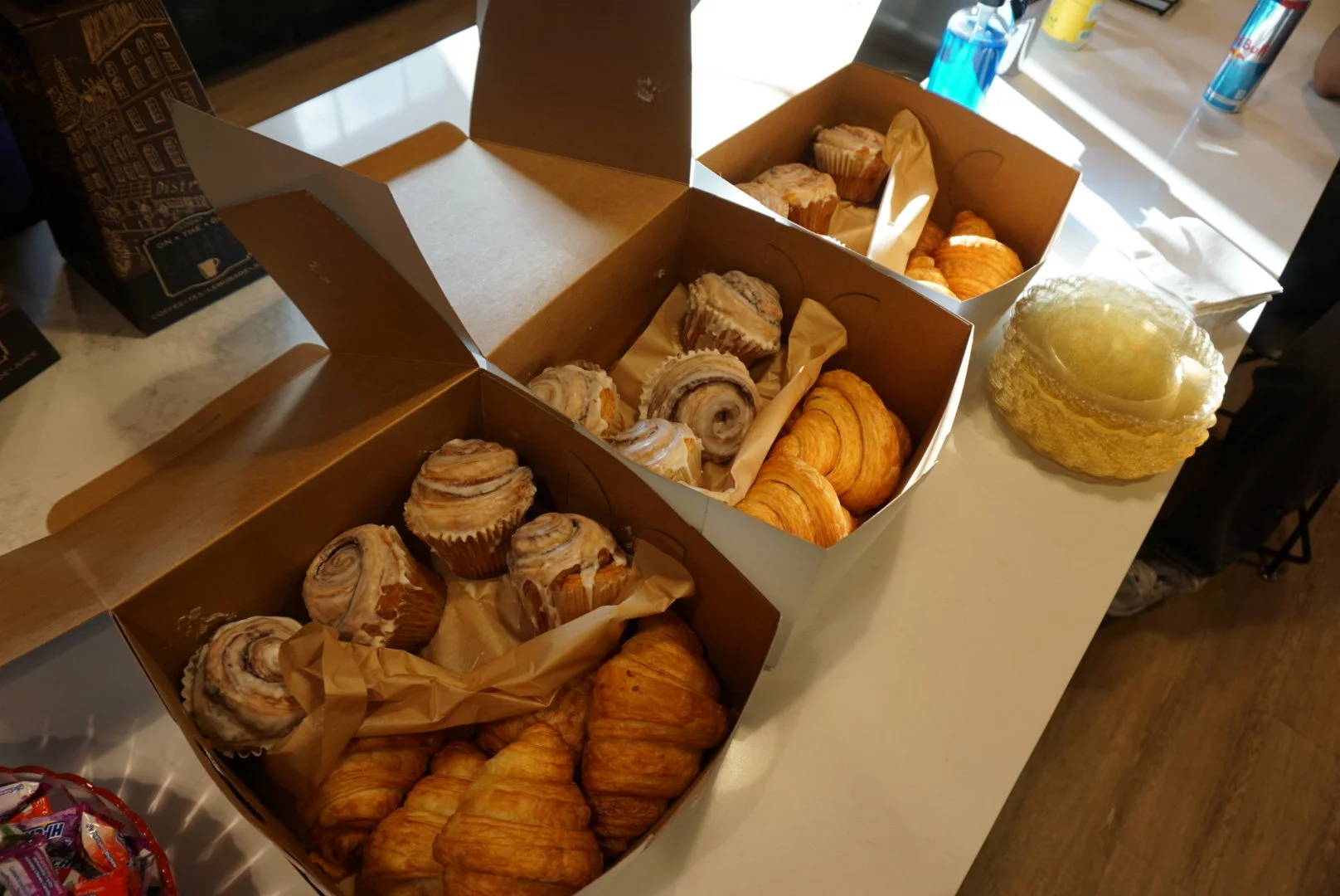  Three cardboard pastry boxes filled with variety of baked pastries next to stack of glittered scalloped transparent plates 