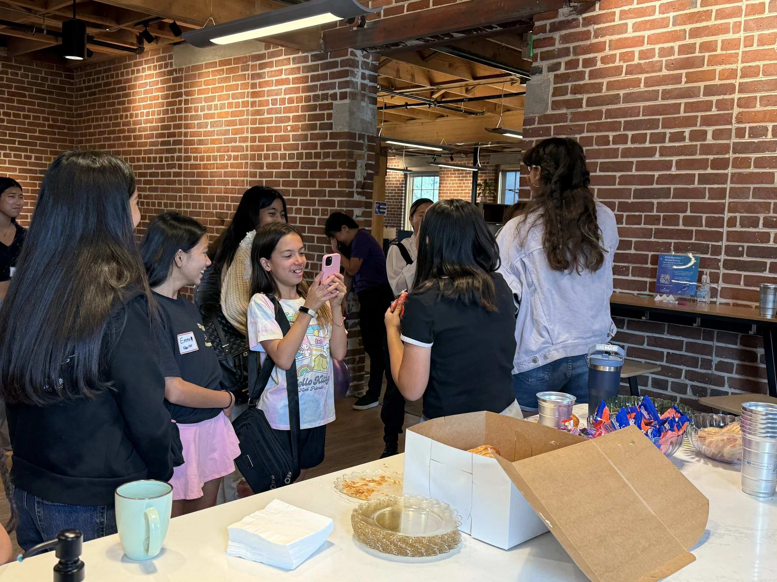  Students in groups conversating standing by kitchen island; room with wooden ceilings, brick walls, decorated with multicolored streamers and balloons; table with white pastry box, napkins, and cups 