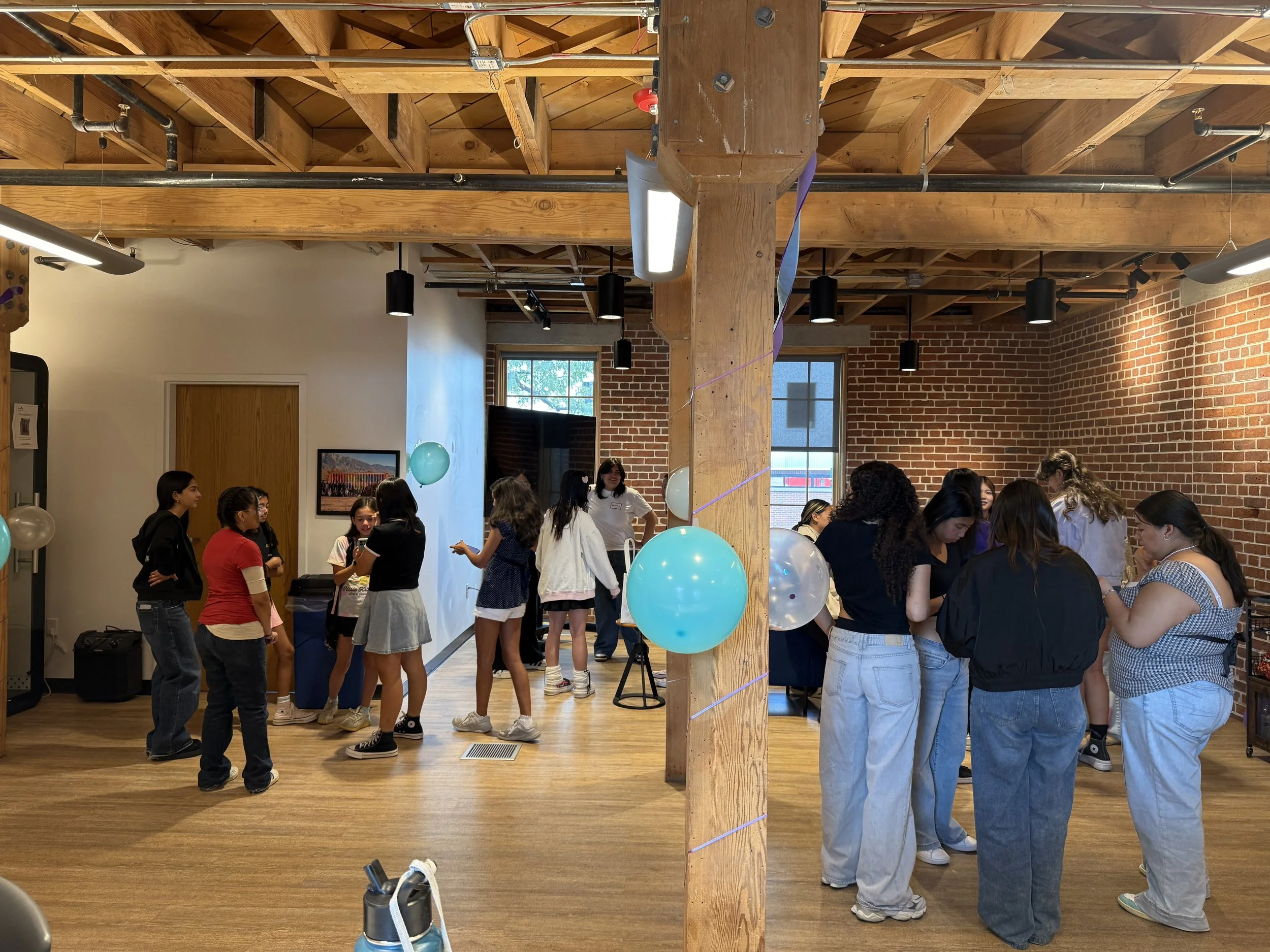  Students in groups conversating standing on wooden floor; room with wooden ceilings, brick walls, decorated with multicolored streamers and balloons 