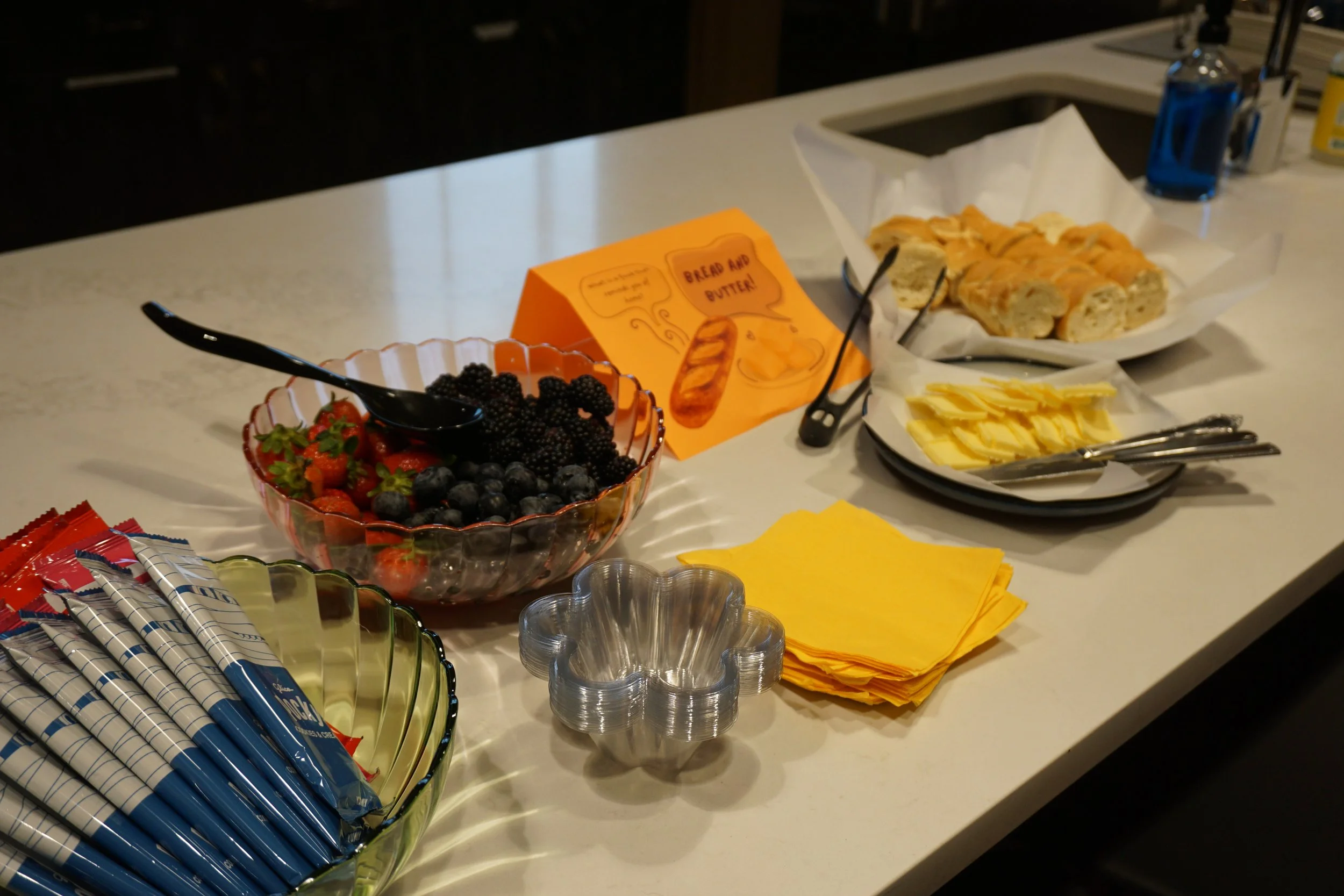  Bowl of berries in a clear scalloped bowl next to other colorful bowls of snacks; yellow napkins next to a plate of sliced bread and butter on a kitchen island 