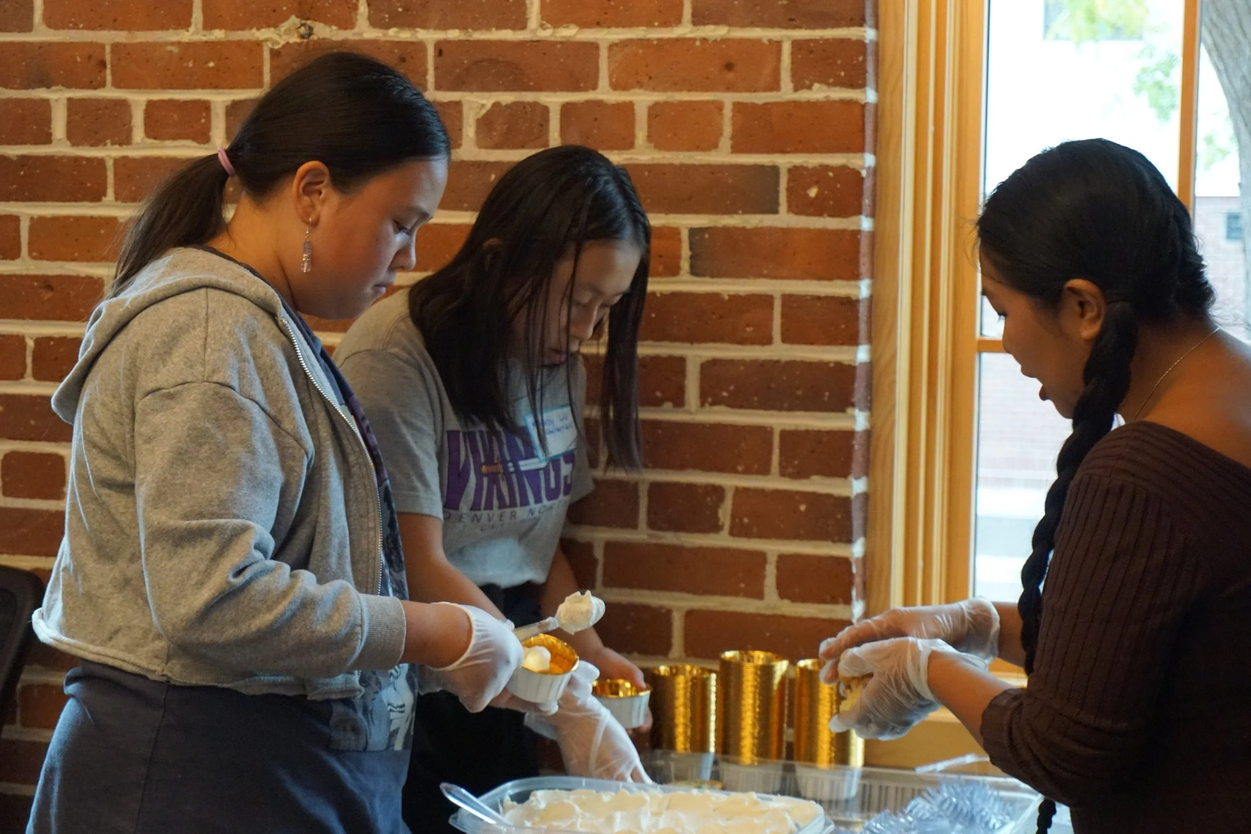 Students with gloves look at icing, cake, and sprinkles; in a classroom with brick walls; vertical windows and shades, with Thoa Nguyen demonstrating 