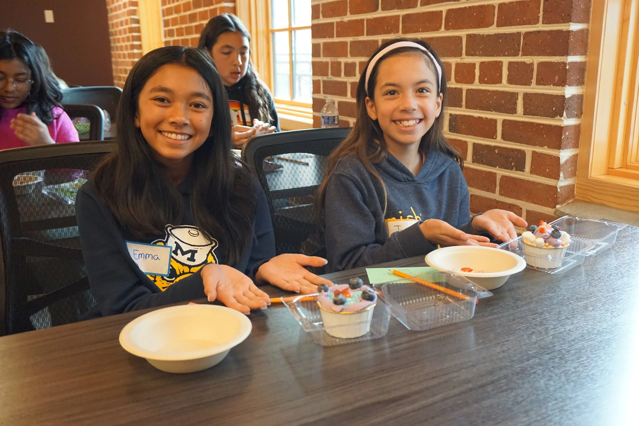  Students pose with cake cups with icing, cake, and sprinkles; in a classroom with brick walls; vertical windows and shades 