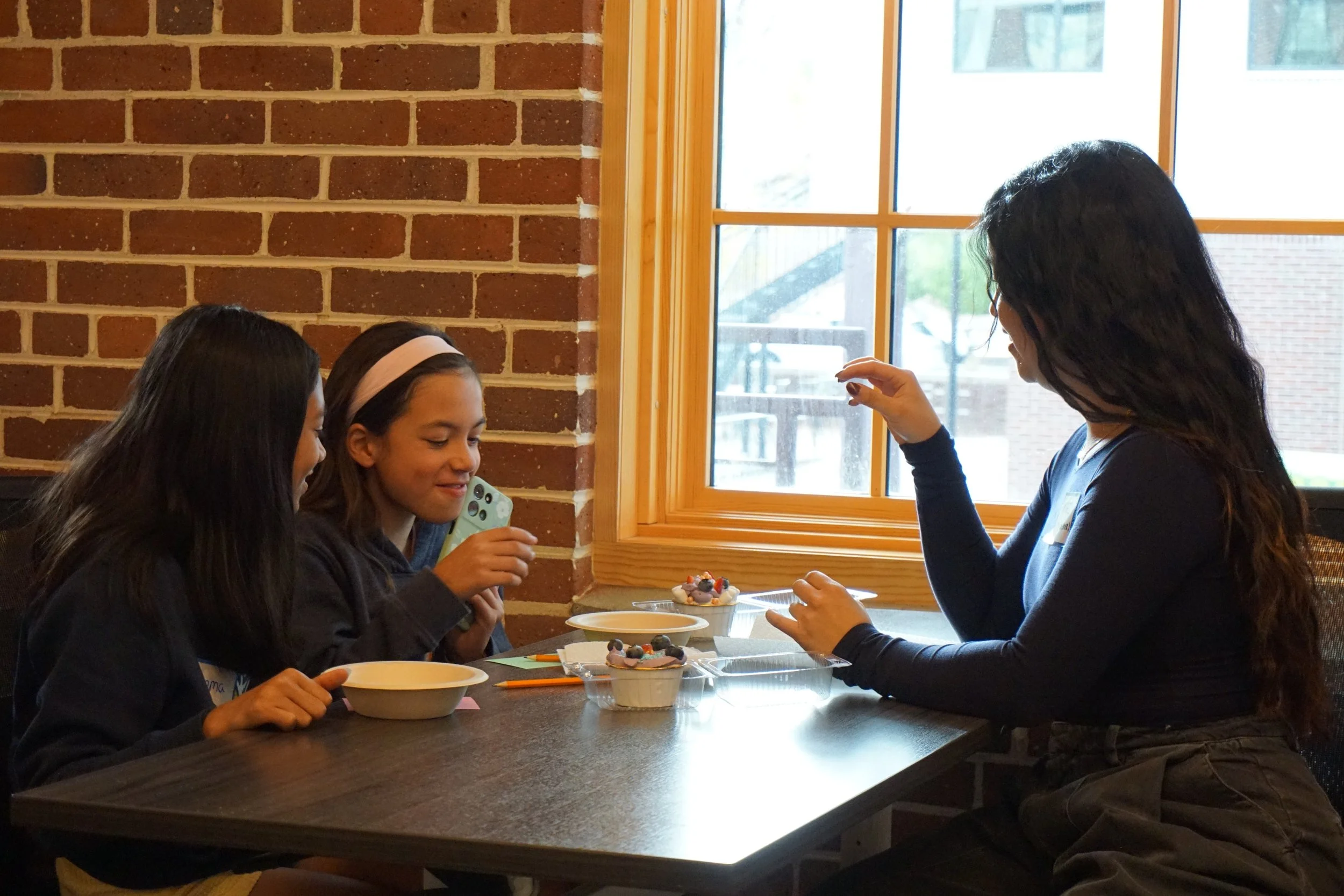  Students make cake cups with icing, cake, and sprinkles with Annabel Kaey; in a classroom with brick walls; vertical windows and shades, against a plum colored wall with a TV 