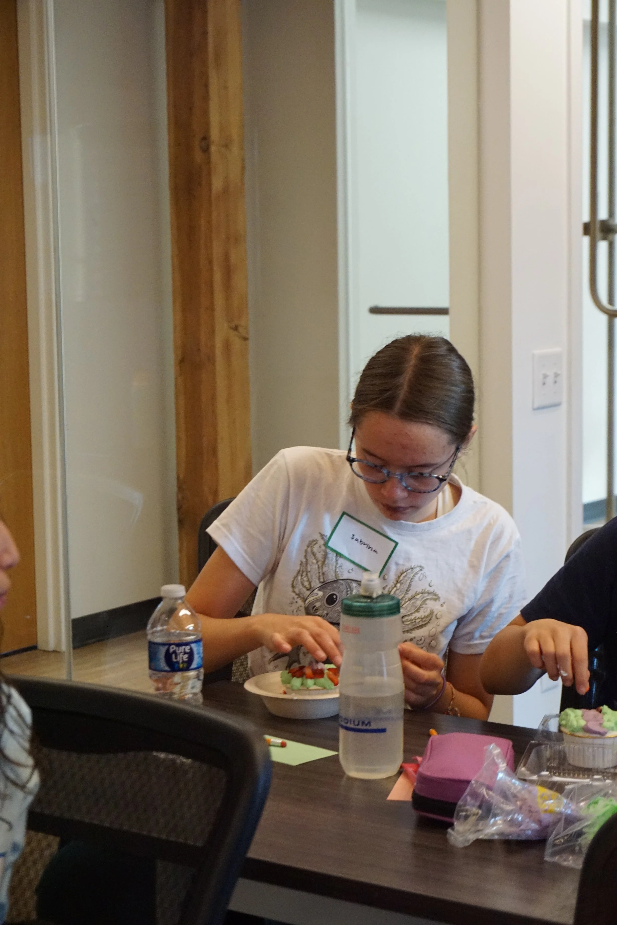  Student makes a cake cup with icing, cake, and sprinkles; in a classroom with glass walls 