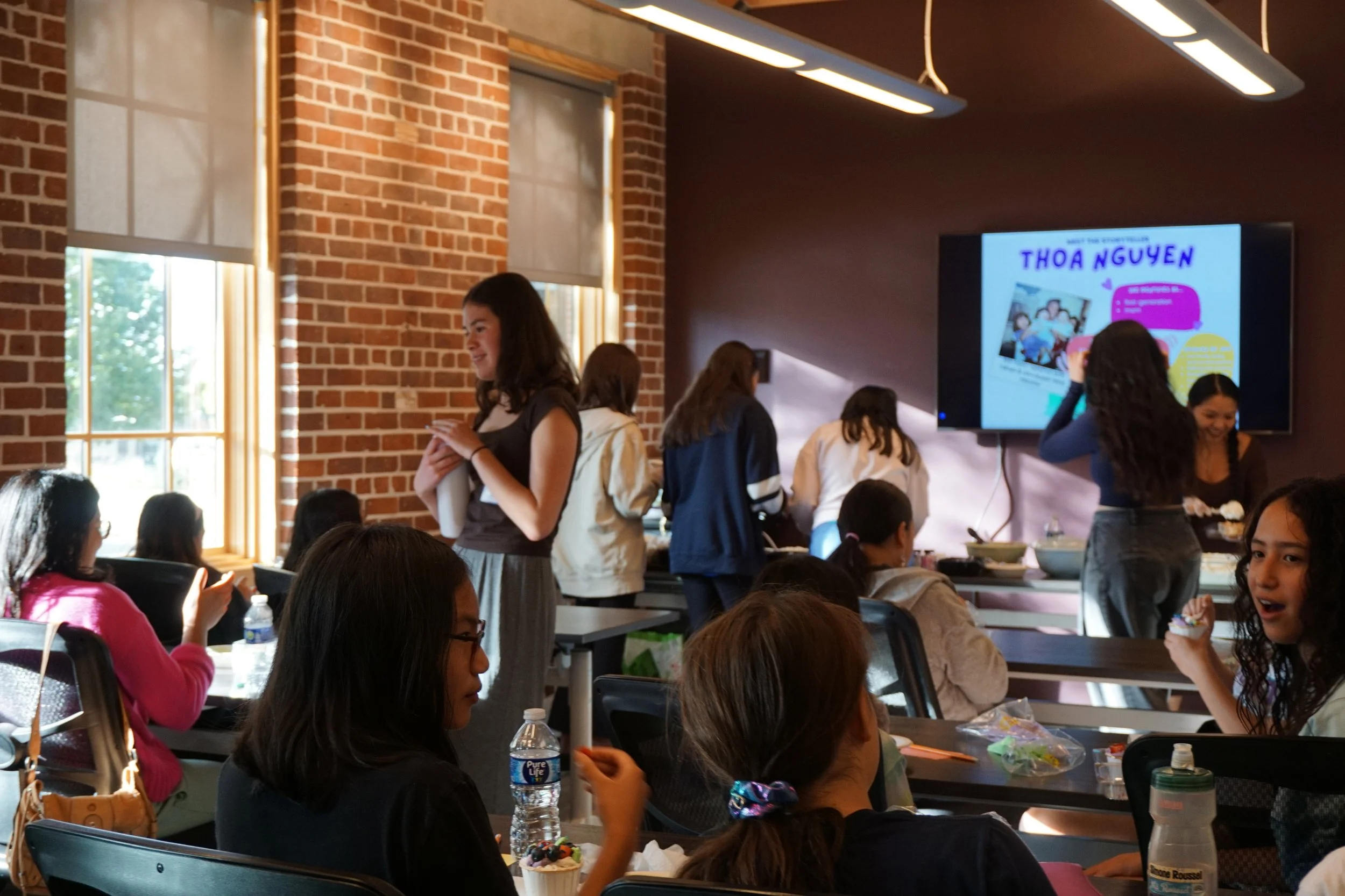  Students in a classroom with brick walls; vertical windows and shades, against a plum colored wall with a TV 