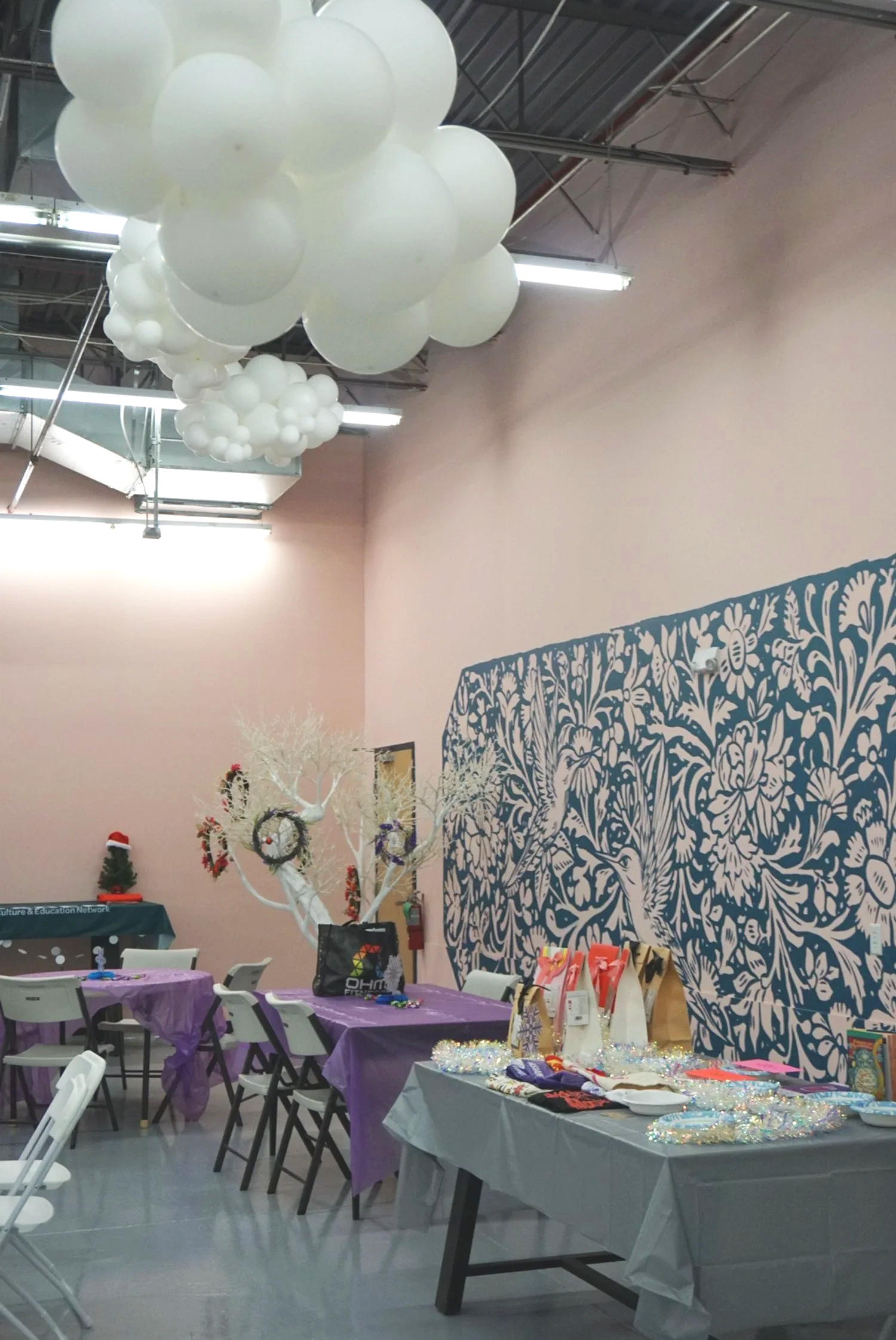  Photo of AGI Holiday Social crafting table at CACEN’s Social Fabric Hub with mural on right wall and balloon clouds hanging from ceiling 