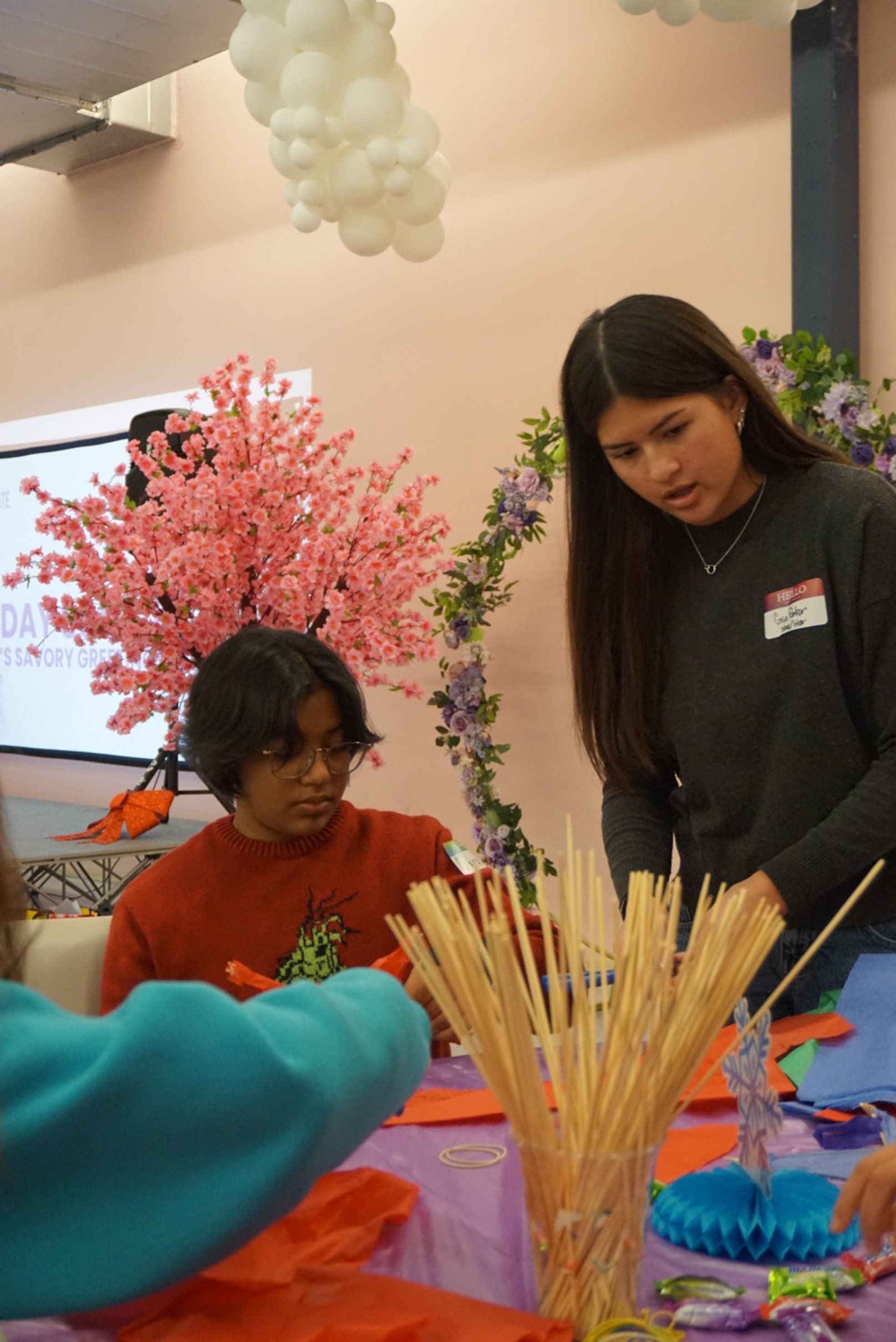  AGI student sitting and standing to create Filipino Parol at AGI Holiday Social; faux cherry blossom tree in the back next to a floral arch.  